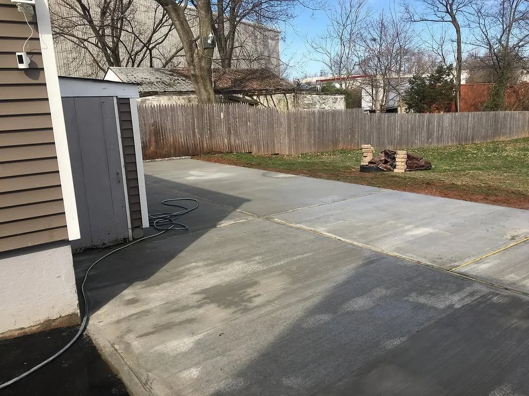 Newly poured concrete driveway with a hose on it, adjacent to a house with beige siding, a wooden fence, and a backyard with patches of grass and a pile of bricks and a tarp.