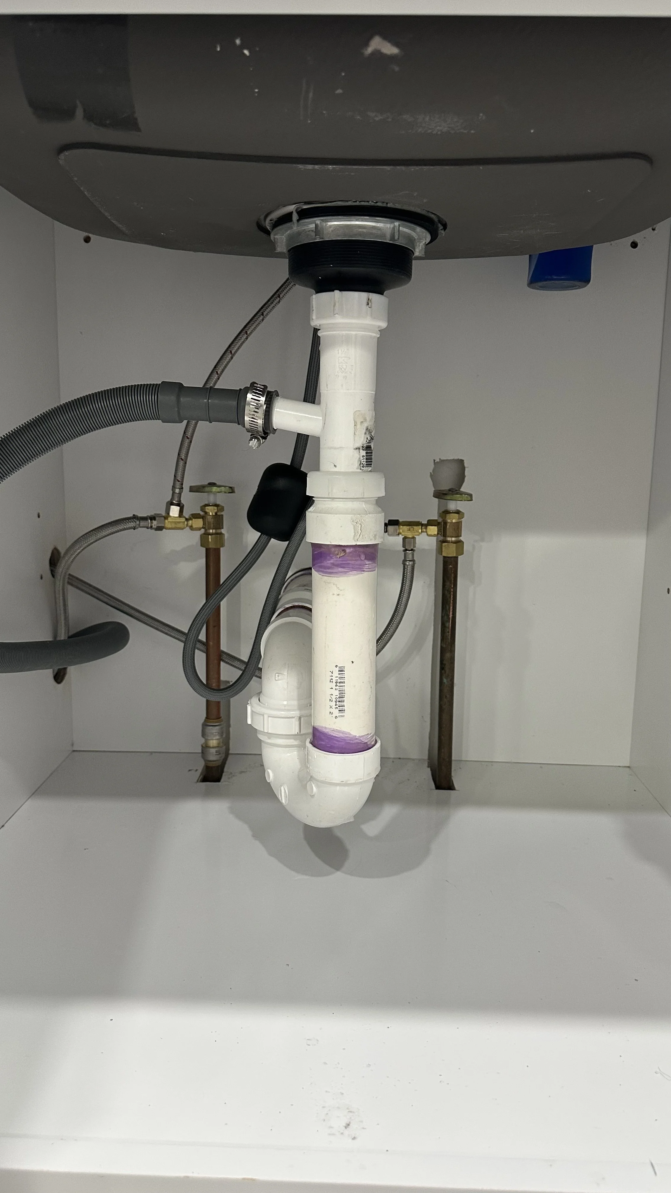 Underneath a kitchen sink showing plumbing pipes, flexible hoses, and shut-off valves.