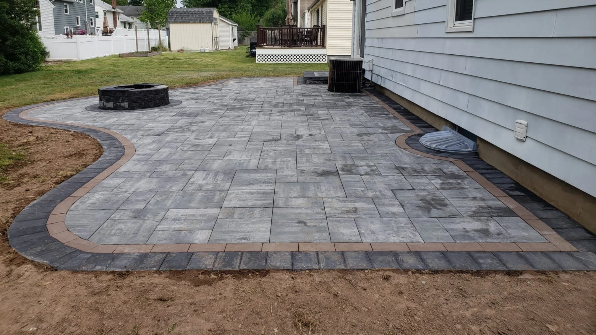 A backyard patio with gray and brown pavers, a fire pit, and a white house with gray siding.