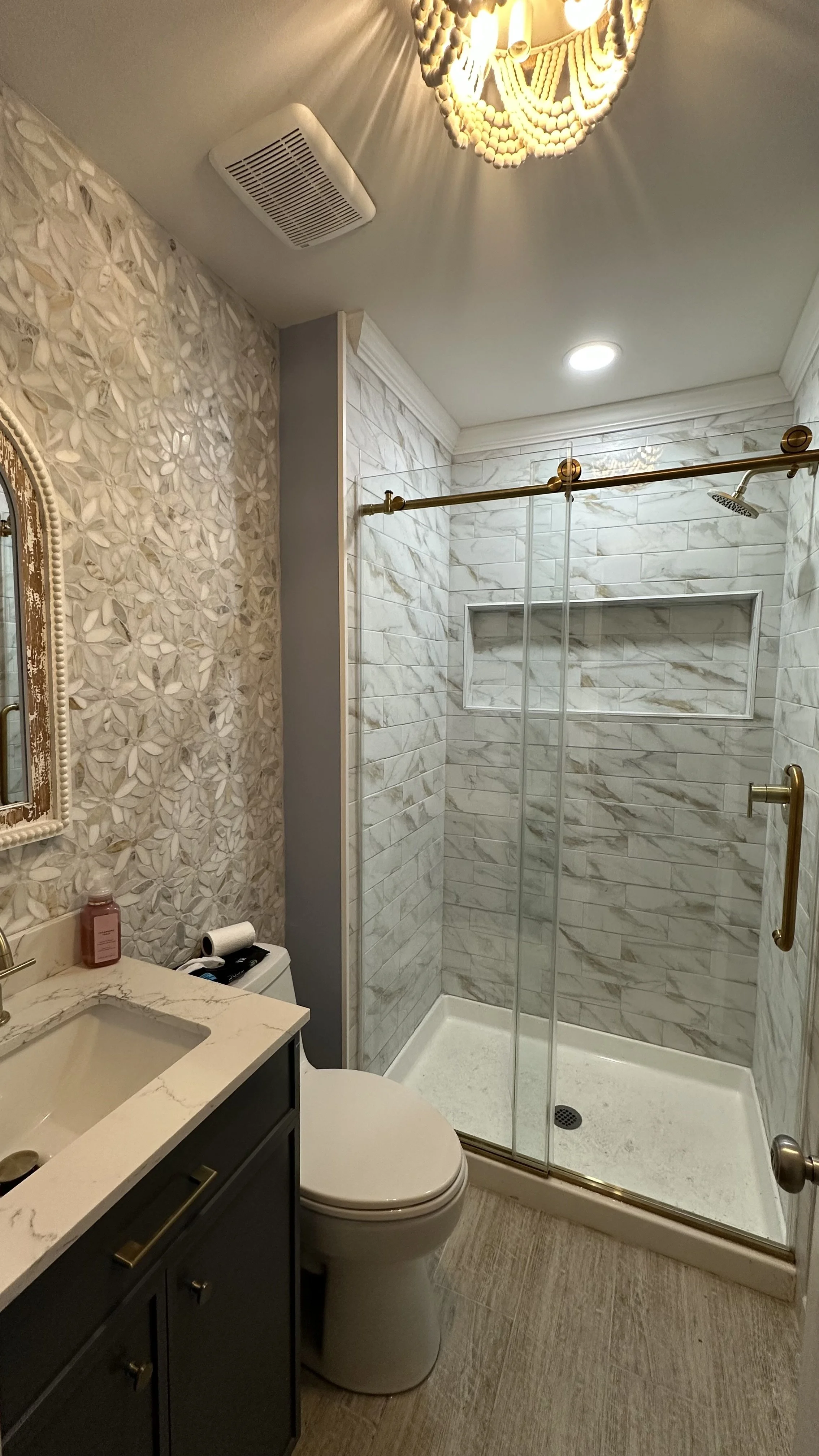 A small bathroom with a patterned wallpaper wall, a white marble countertop vanity with dark cabinets, a toilet, and a glass-enclosed shower with marble tile walls. There is a decorative mirror and a light fixture on the ceiling.