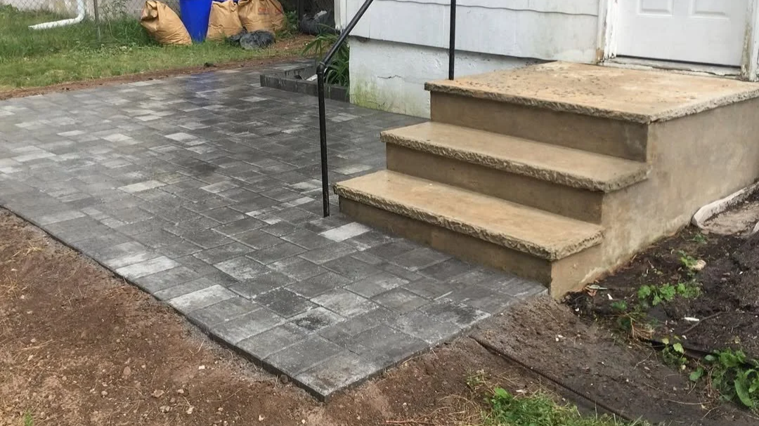 Newly paved concrete and brick stairs leading to a door, with a small ramp for accessibility and a black handrail, next to a patch of dirt and grass.