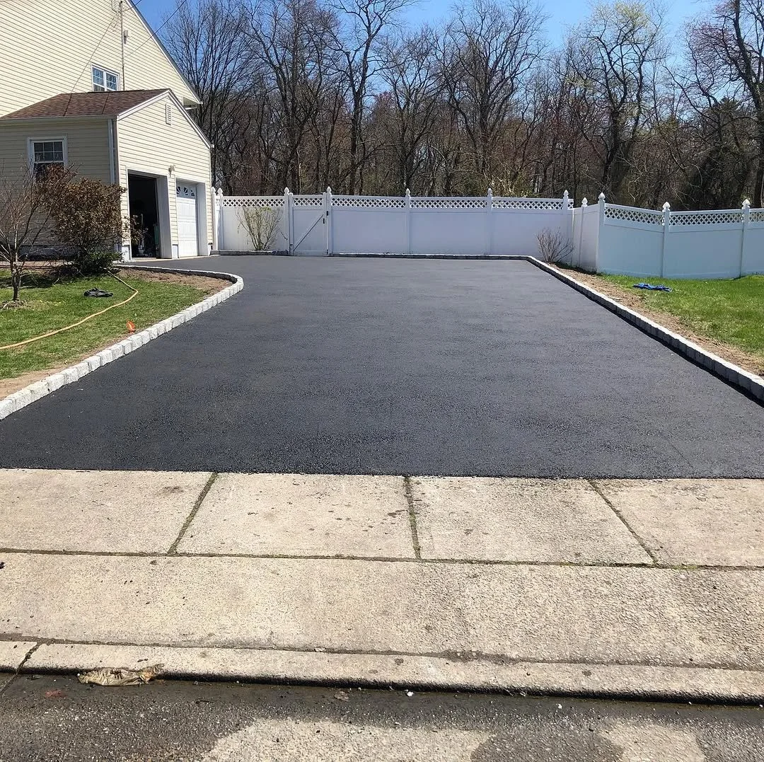 Freshly paved driveway in front of a house, bordered by light gray pavers, with a white vinyl fence in the background, and a grassy yard on either side.