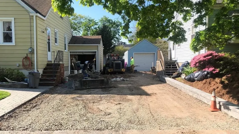 Residential driveway under construction with workers, machinery, and construction materials, surrounded by houses and greenery.
