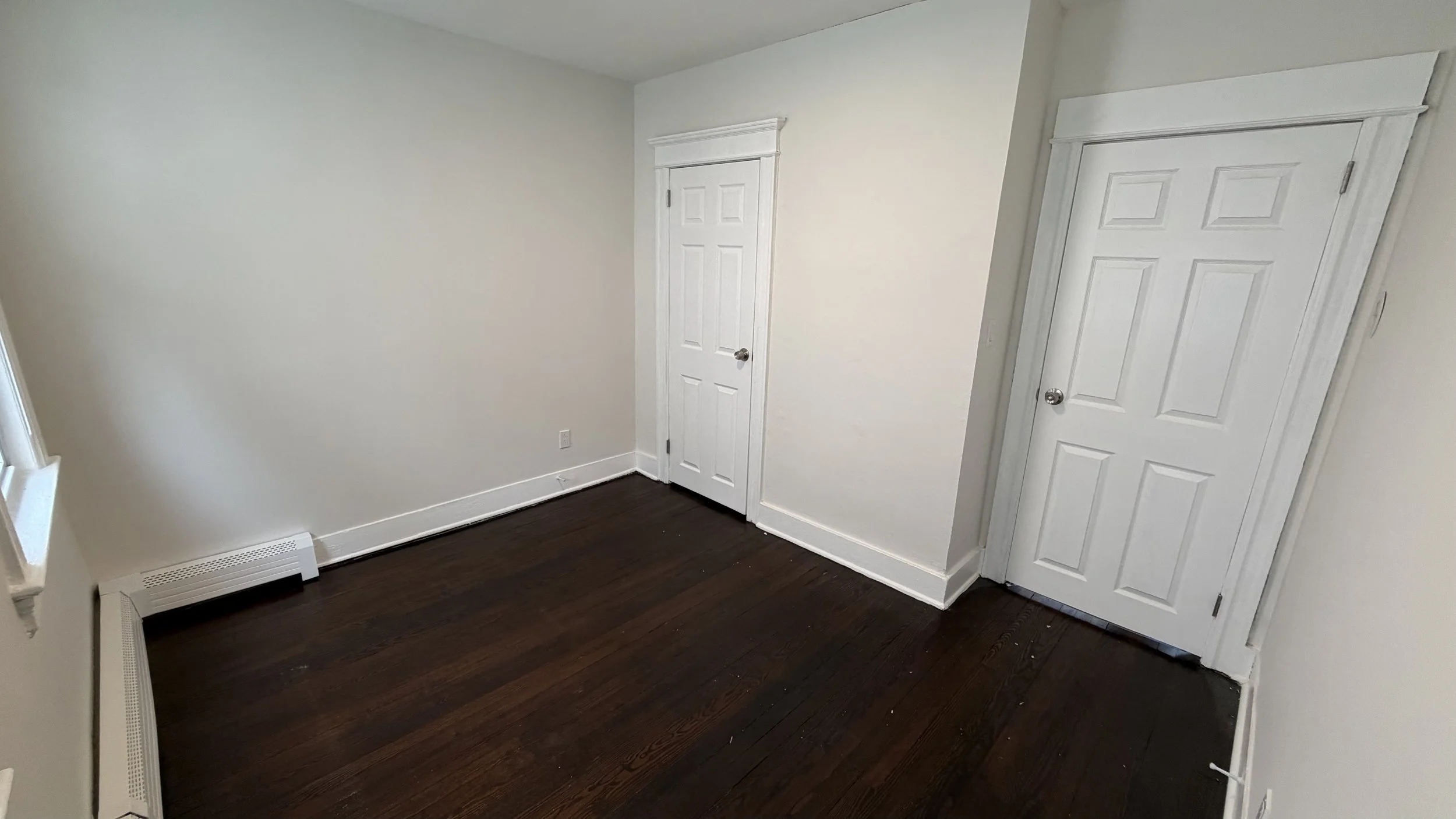 Empty room with white walls, dark hardwood floors, two closed white doors, and a window on the left side.
