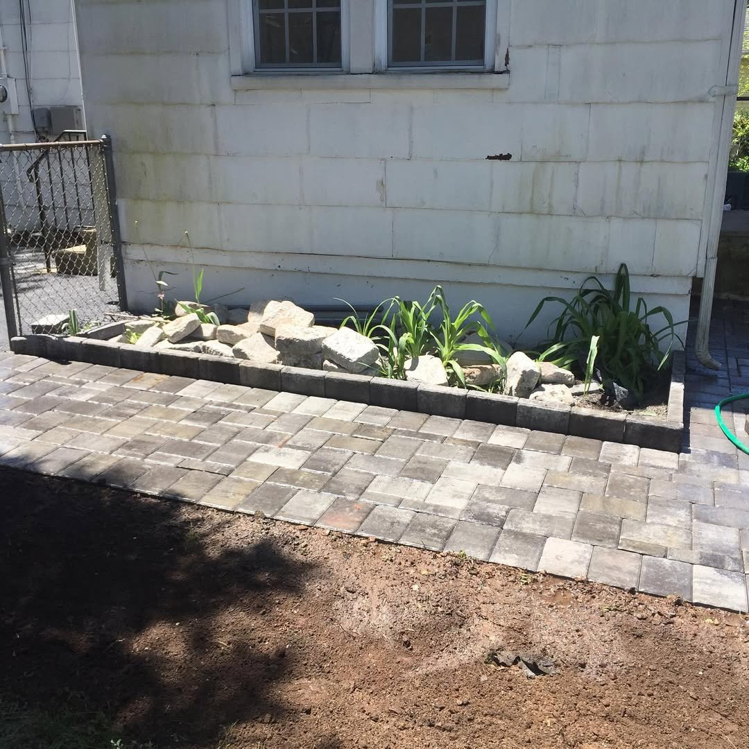 A small garden bed with rocks and green plants in front of a white house wall, next to a new paved pathway.