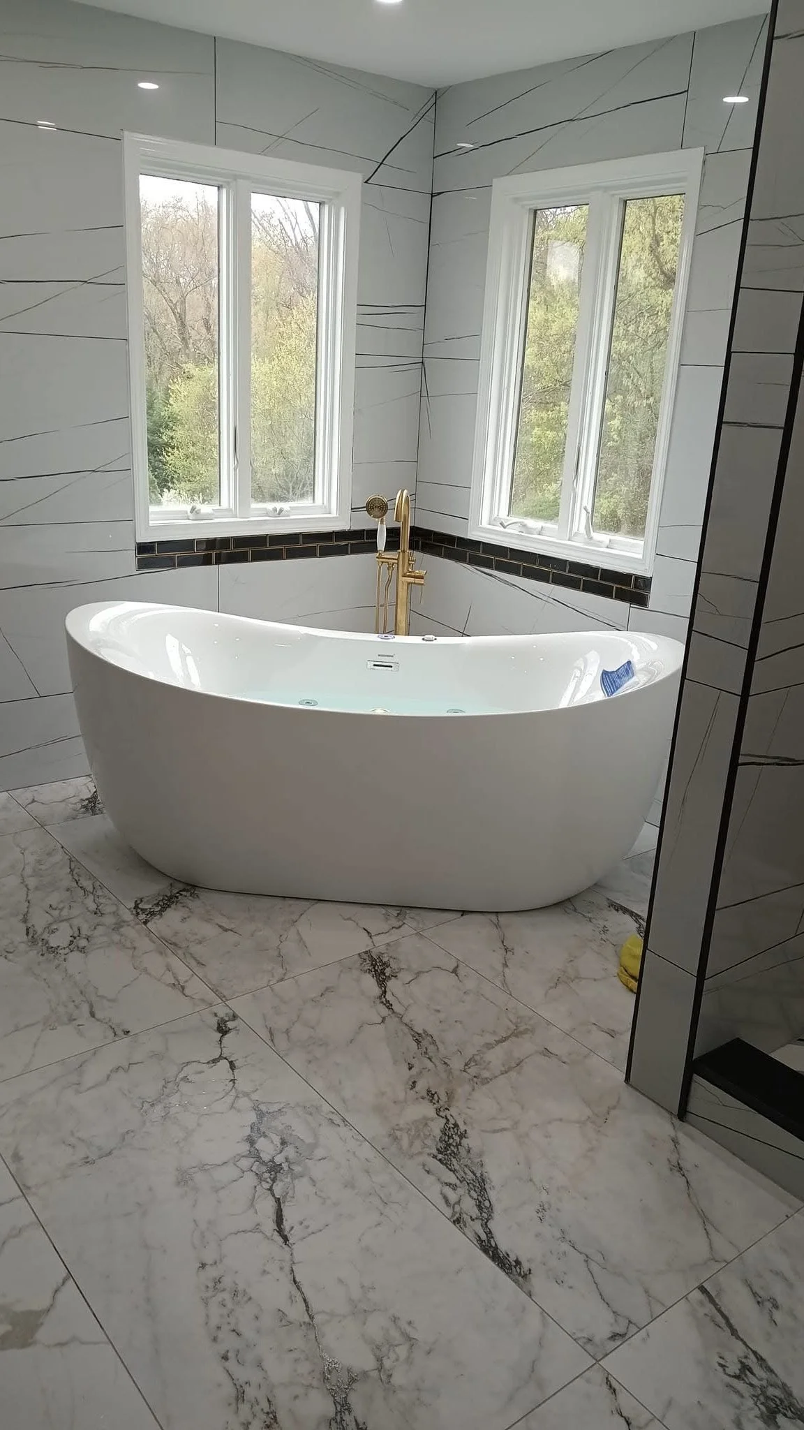 A modern bathroom with a white freestanding bathtub placed near two large windows, black tile trim, marble-like floor tiles, and white walls with black veining, equipped with a gold faucet and fixtures.