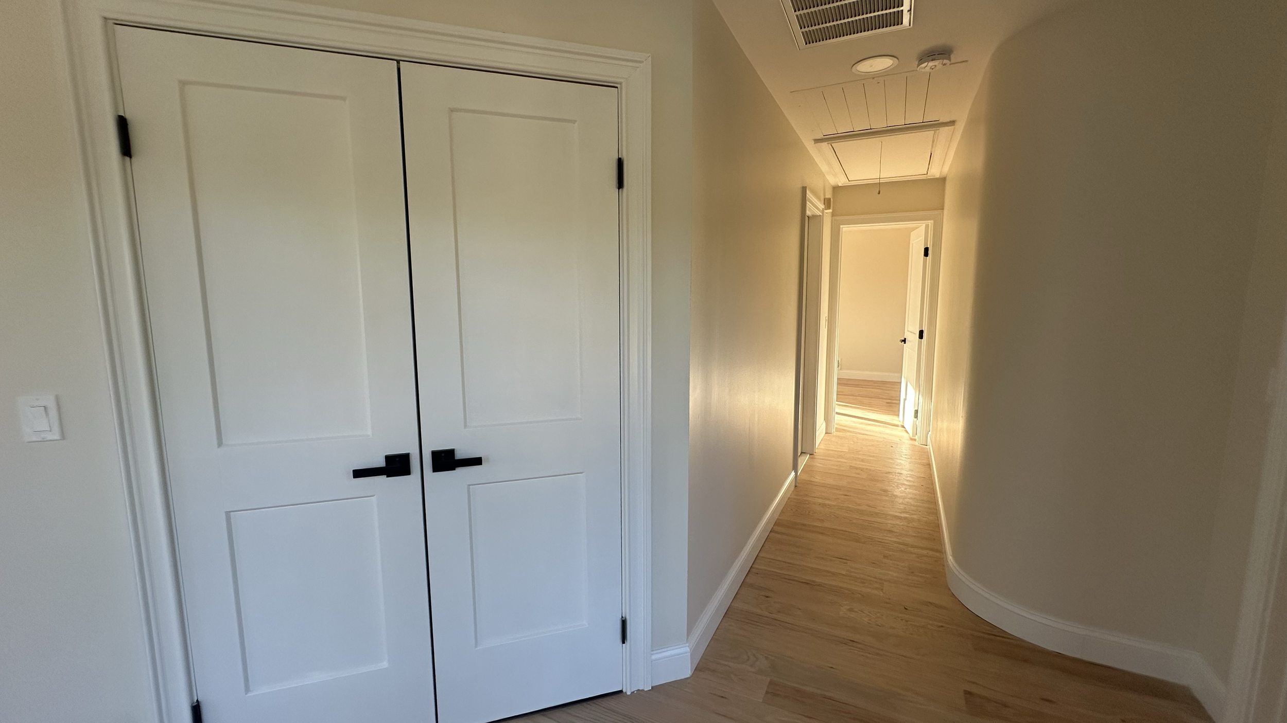 An empty hallway with light-colored walls and wooden flooring, featuring a closed closet with white double doors and black handles on the left, and a door at the end of the hallway.