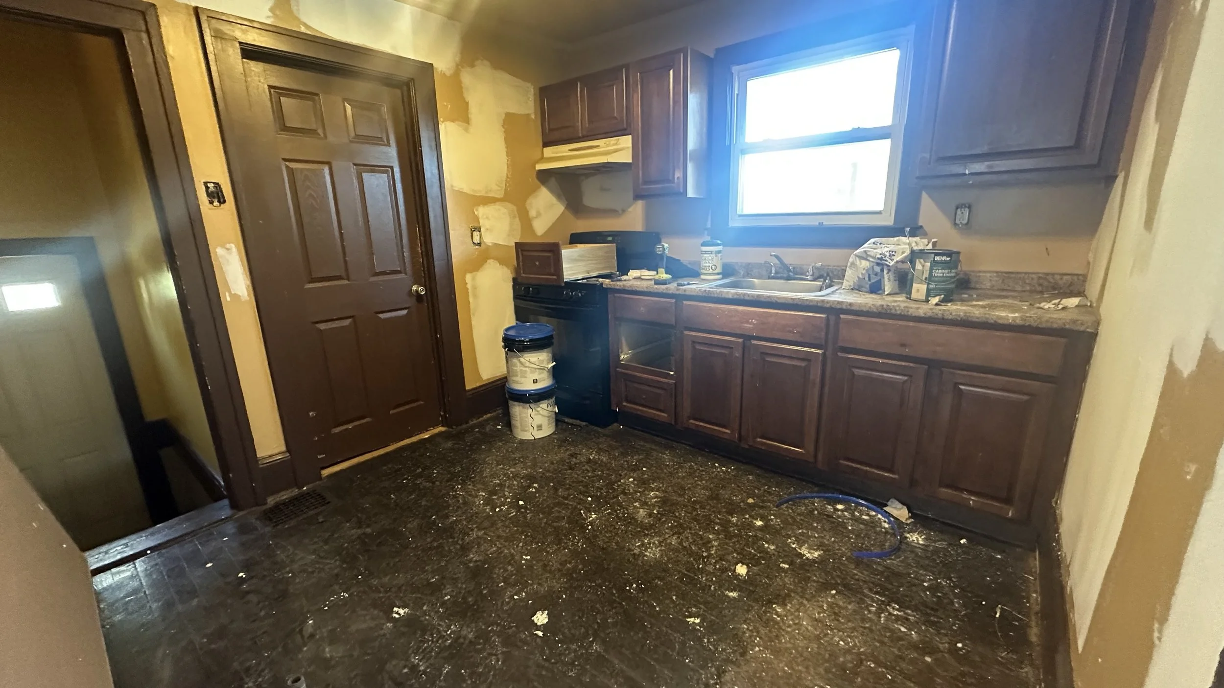 Kitchen in the process of renovation with unfinished yellow walls, dark wooden cabinets, a window above the sink, and a black stove. The floor is dirty with debris, and paint cans are stacked near the stove.