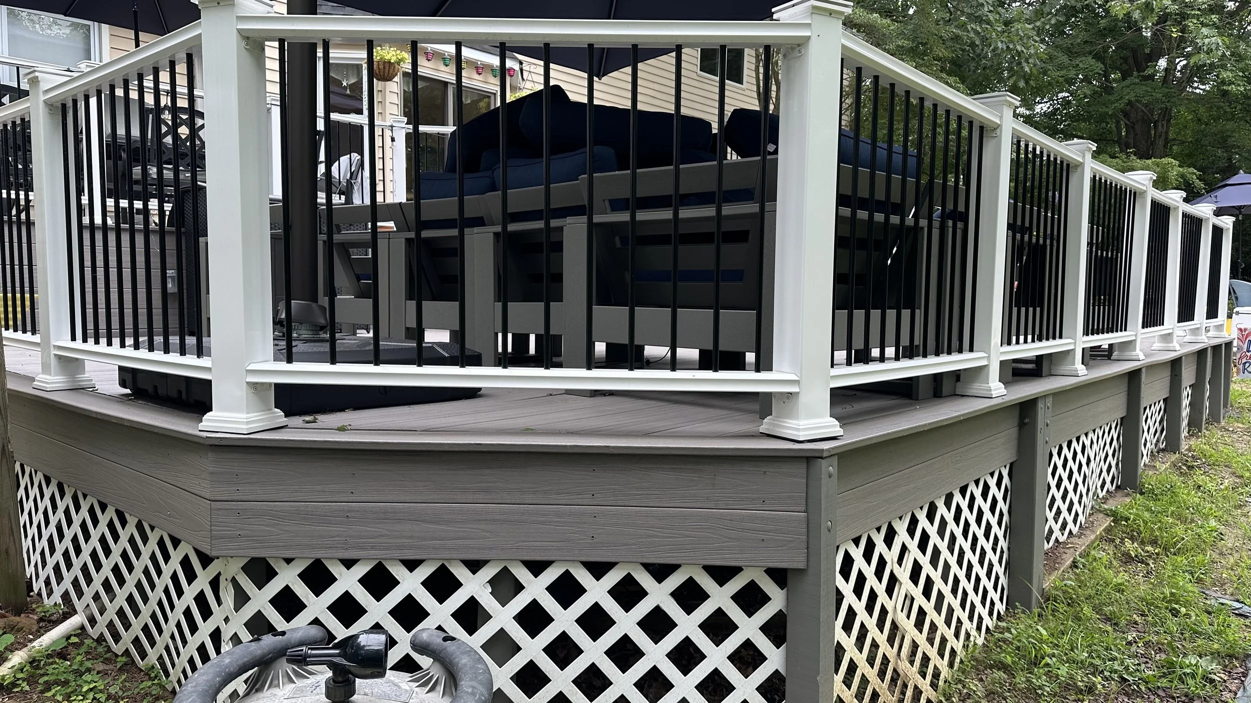 A raised outdoor deck with white and black railing, furniture, and a large black umbrella, surrounded by green trees and grass.