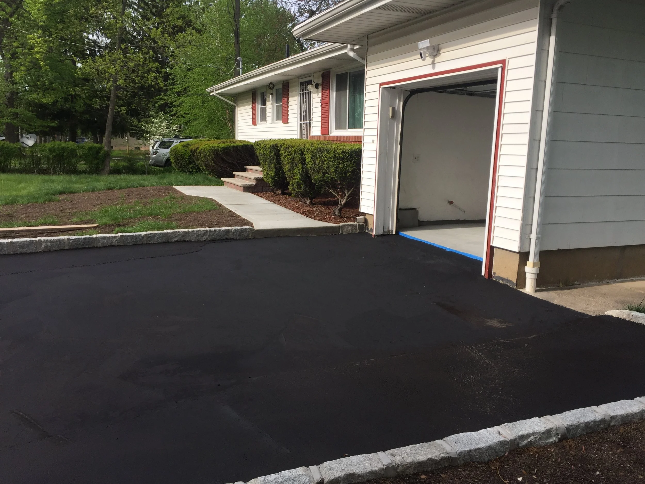 A residential driveway freshly paved in black asphalt with a stone border, leading to an open garage attached to a white house with red trim; some green trees and bushes are visible in the background.