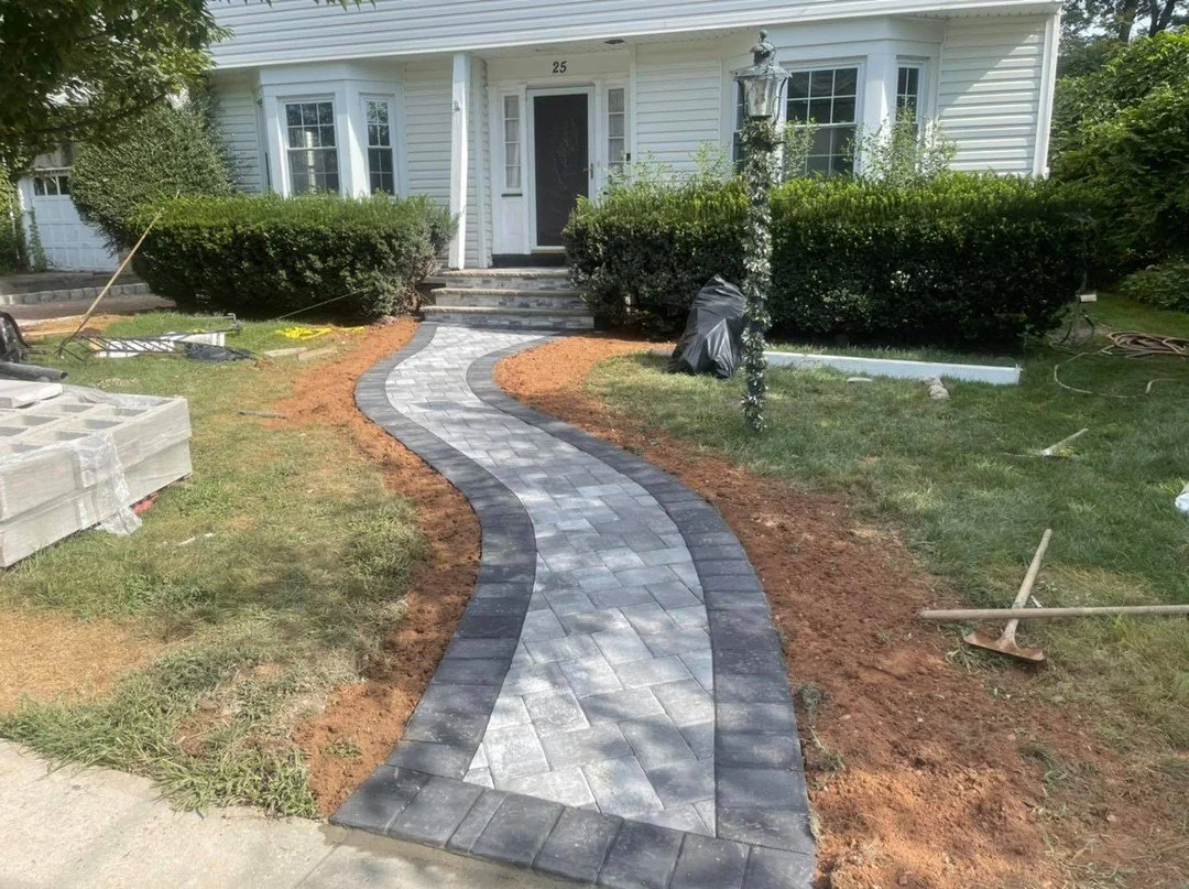 Newly paved winding walkway made of gray and black bricks leading up to a white house with the number 25. Surrounding the walkway are garden beds, shrubbery, and a small patch of grass, with construction tools and materials nearby.