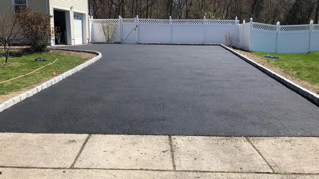 Newly paved black asphalt driveway with beige concrete sidewalk in front, adjacent to a green lawn and white fence in the background.