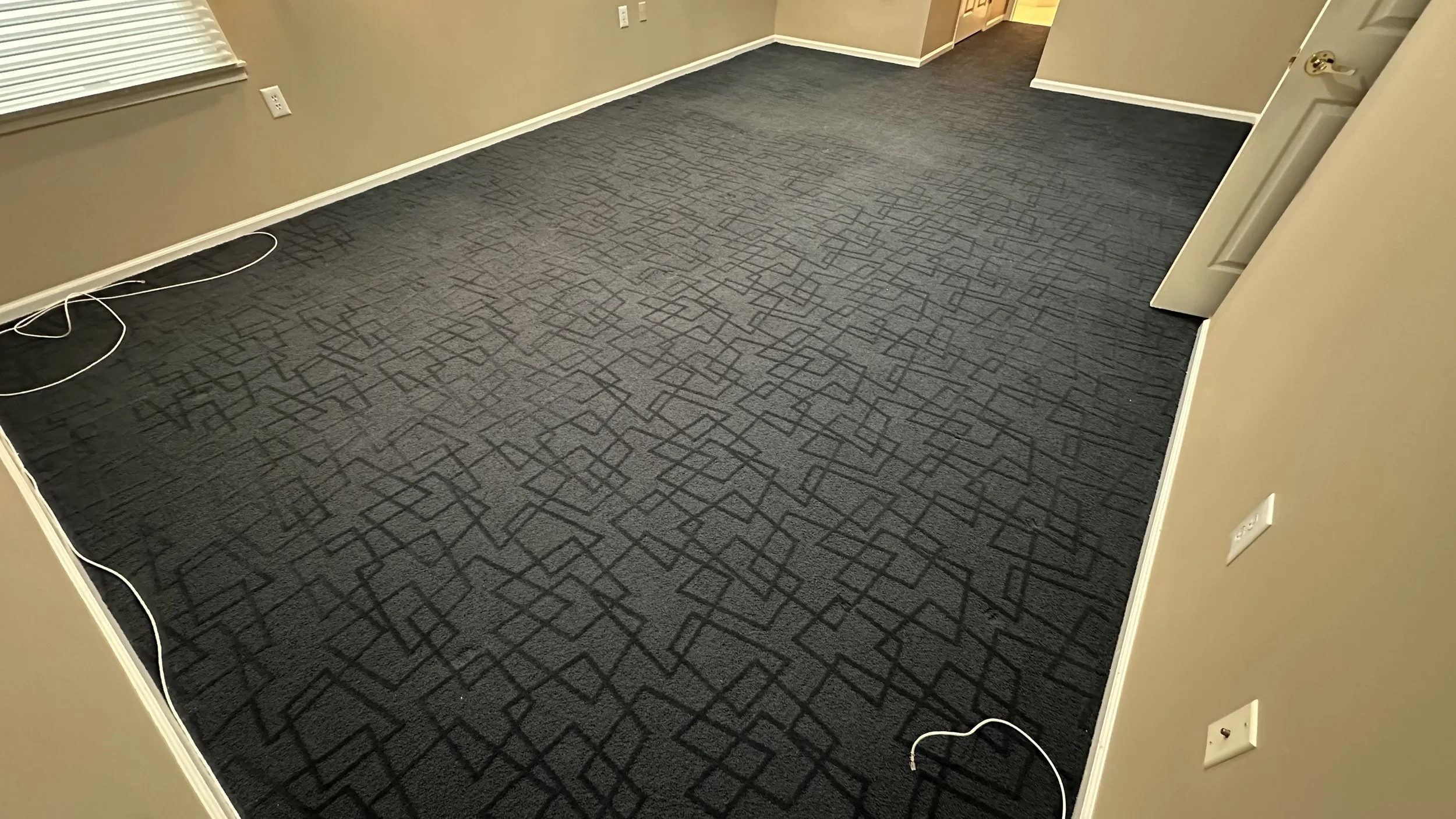 Empty room with beige walls, window with horizontal blinds, black patterned carpet, and electrical outlets on the walls, with white cords on the floor.