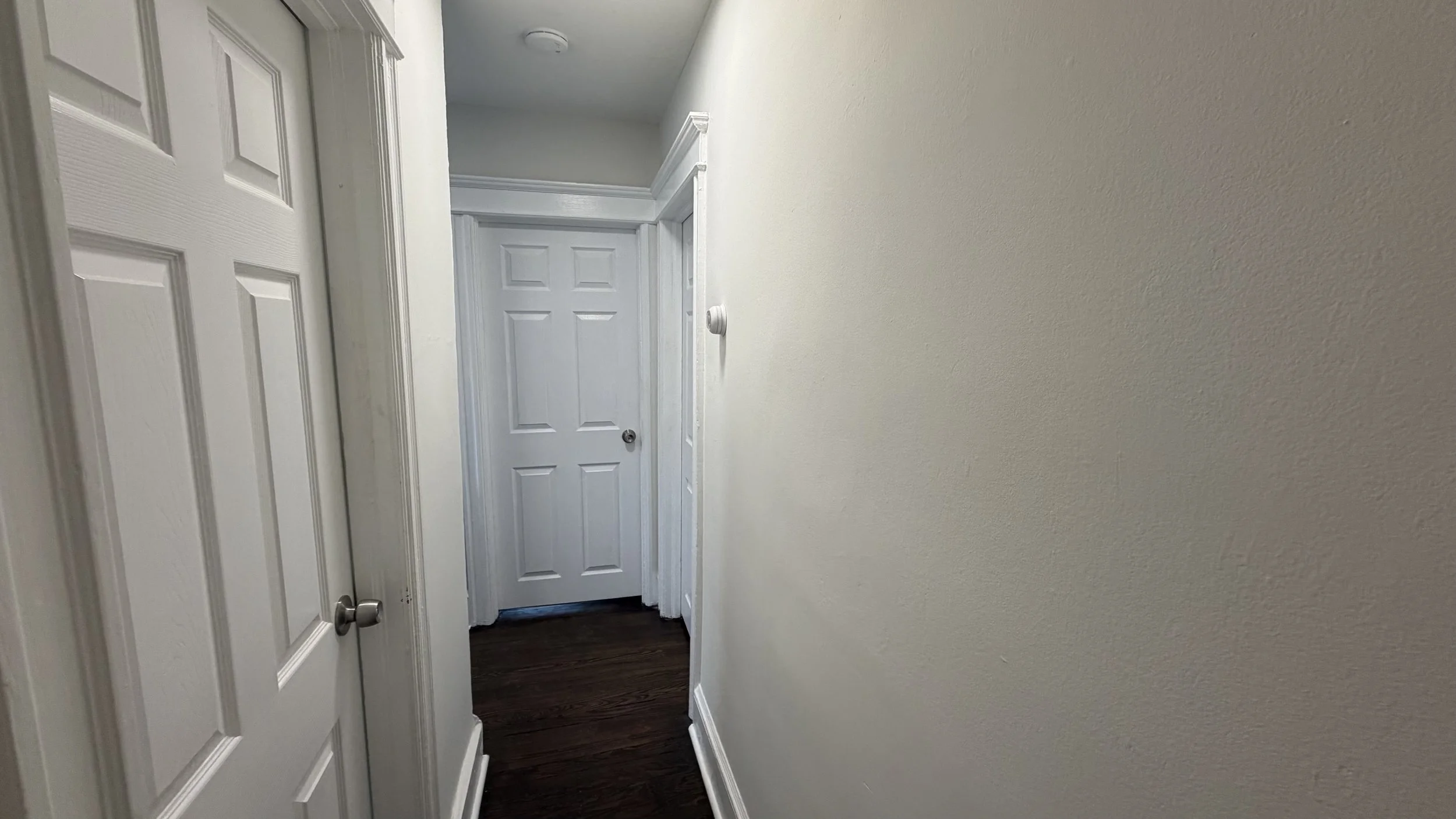 A narrow hallway inside a house with white walls and white doors, and dark wood flooring.