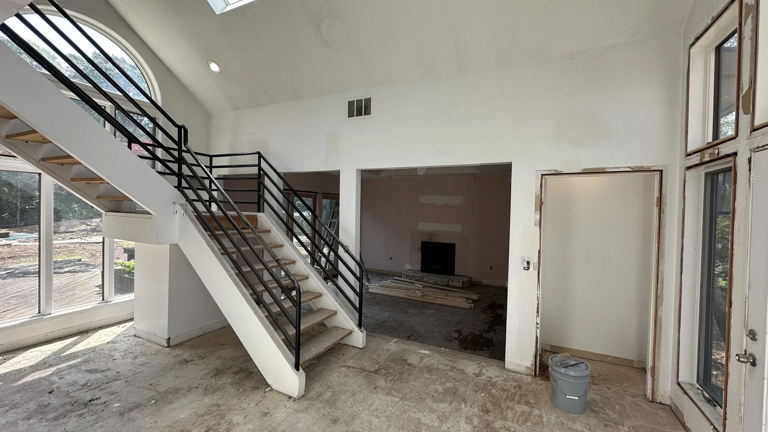 Image of a partially finished living room with an upstairs staircase, large windows, and a fireplace in a house under renovation.