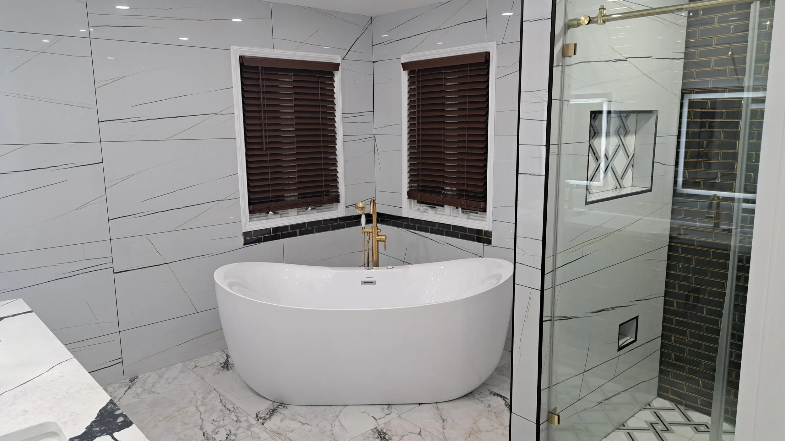A modern bathroom featuring a white freestanding bathtub with a black and gold faucet, marble tiled walls with diagonal veining, brown window blinds, and a glass-enclosed shower with dark brick-style tile accents.