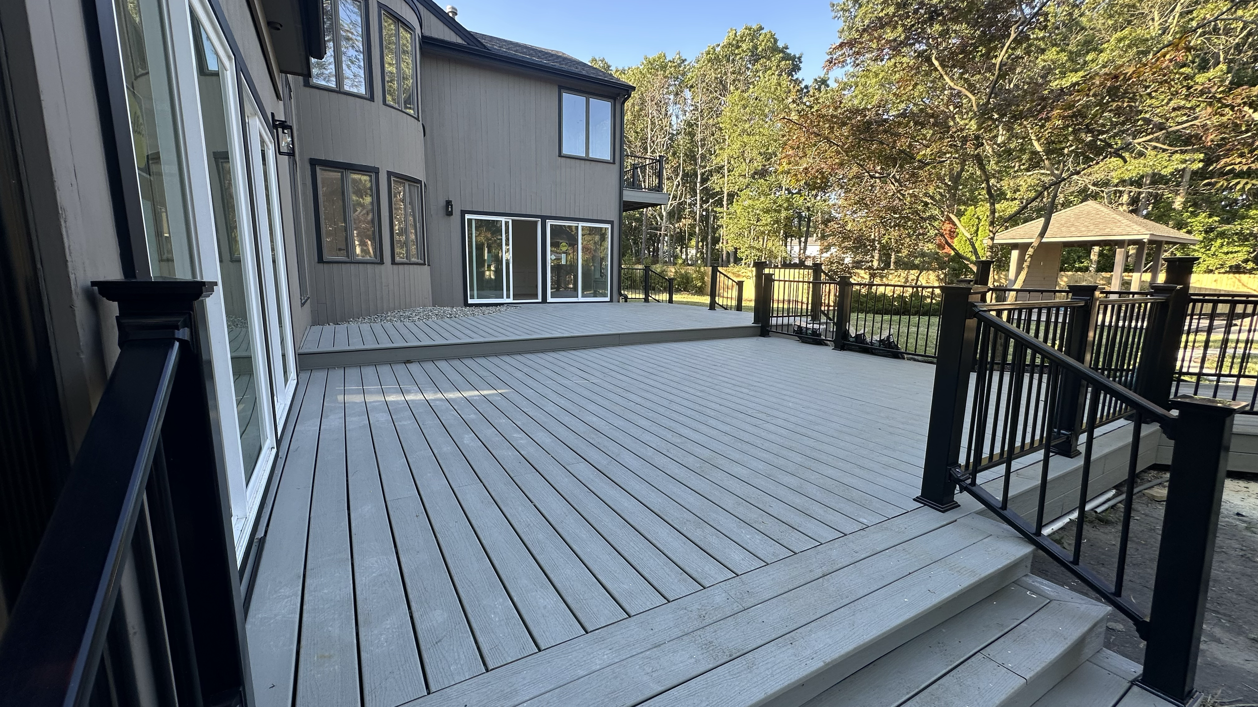 Large wooden deck attached to a gray house with multiple glass doors and windows, surrounded by trees with autumn foliage.