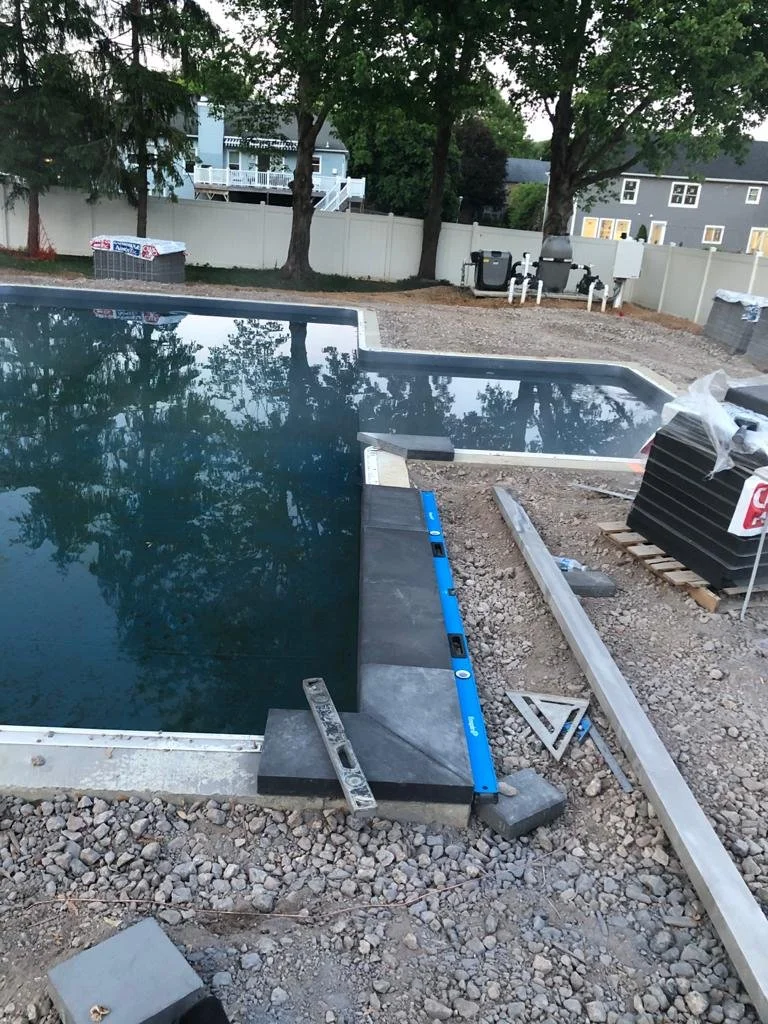 Under construction backyard with a pool, trees, and houses in the background. Pool edges are being finished with tools and materials nearby.