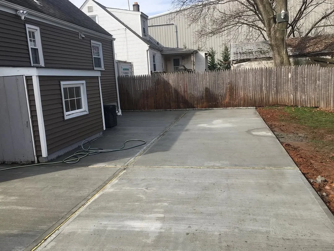 Backyard with concrete patio, brown wooden fence, house with brown siding, some outdoor equipment, and a tree.
