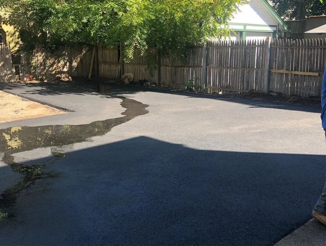 Freshly paved asphalt driveway with a small water puddle reflecting the sky, enclosed by a wooden fence, trees, and a neighboring house with a green roof.