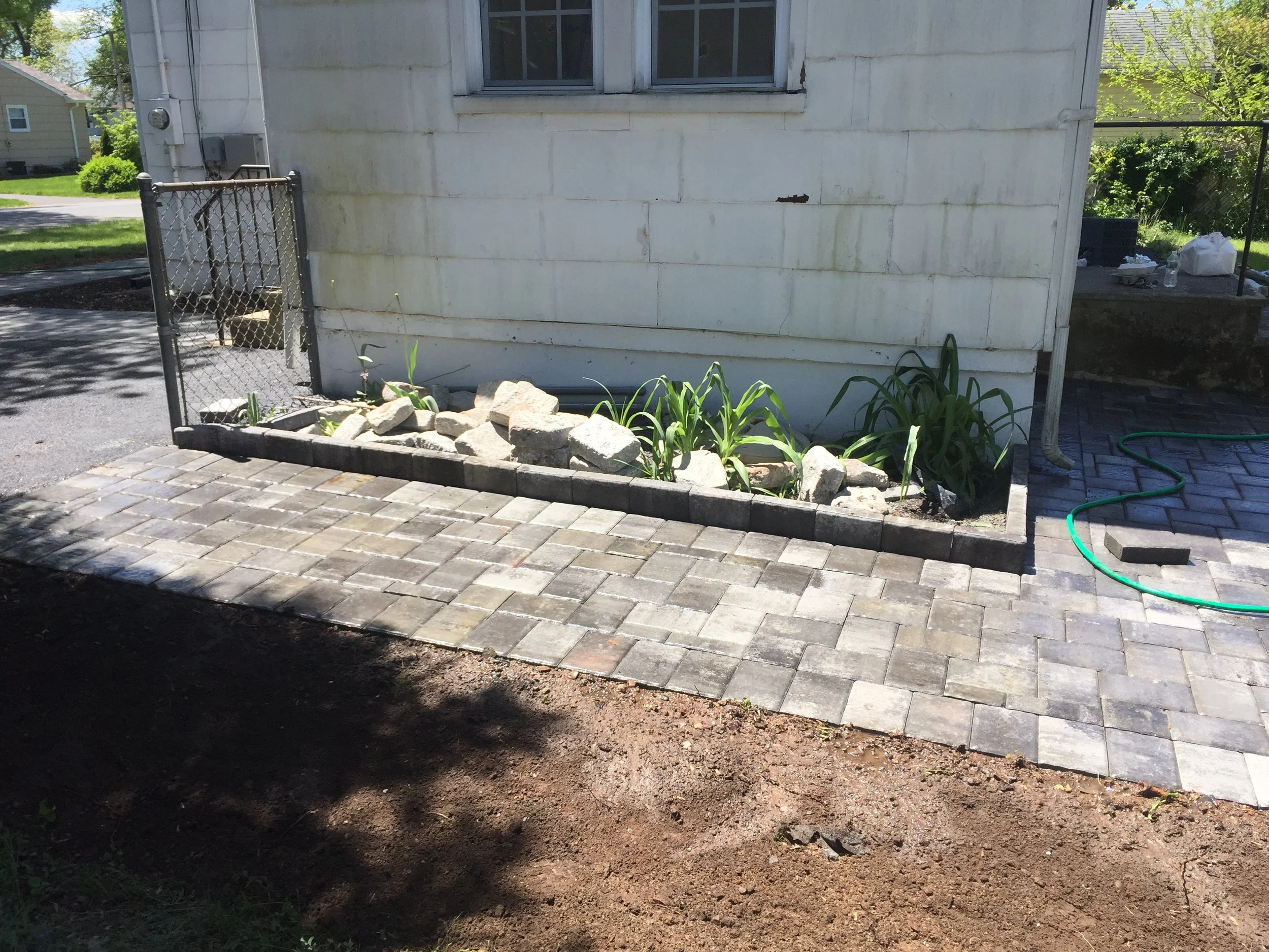 A newly paved brick patio beside a small garden bed with rocks and plants next to a white house with windows.