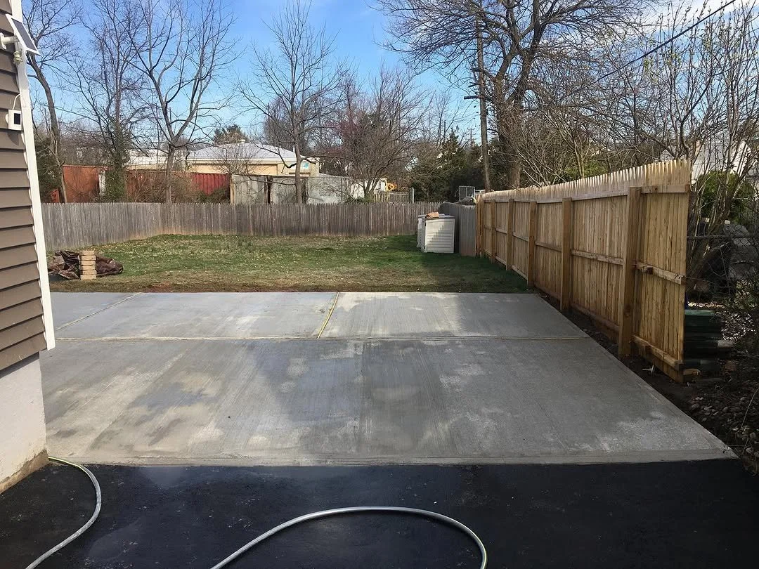 View of a backyard with a concrete patio, a grassy area, a wooden fence, and trees with no leaves.