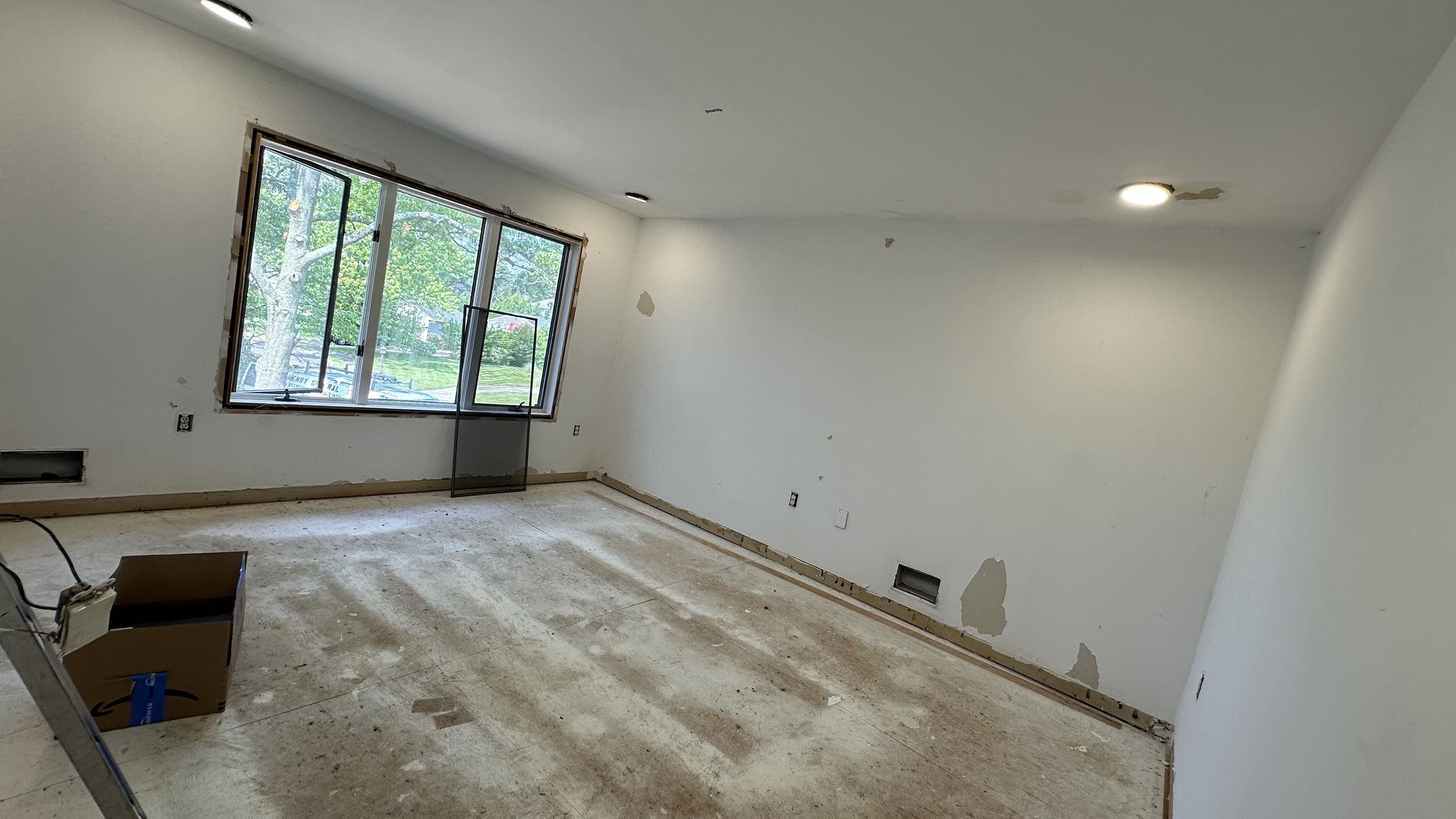 Empty room under renovation with damaged white walls, a large window showing greenery outside, and a floor with construction debris and a cardboard box.