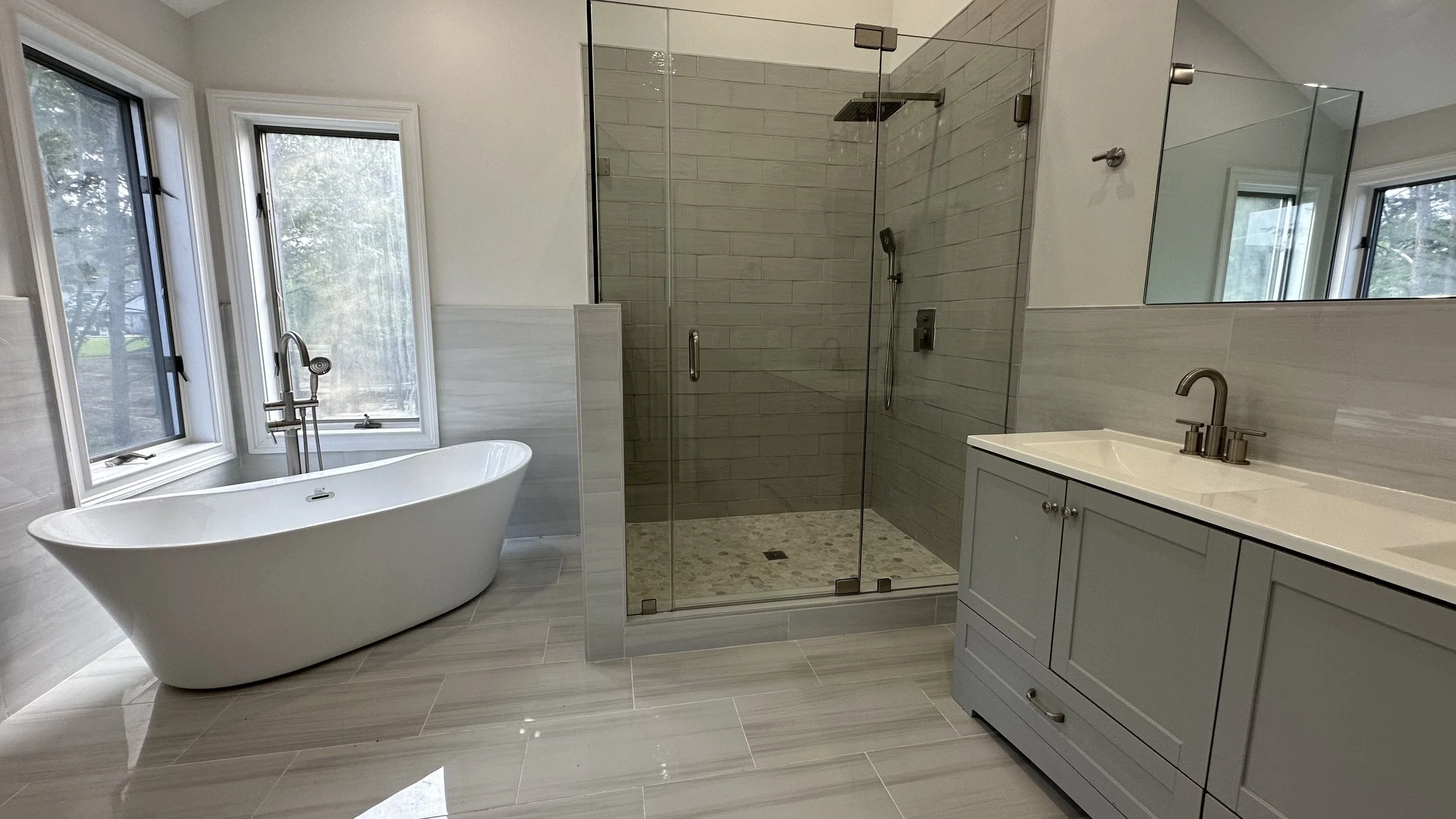 Modern bathroom with a freestanding bathtub, a glass-enclosed shower, and a vanity with a mirror. The space has large windows and neutral-colored tiles.