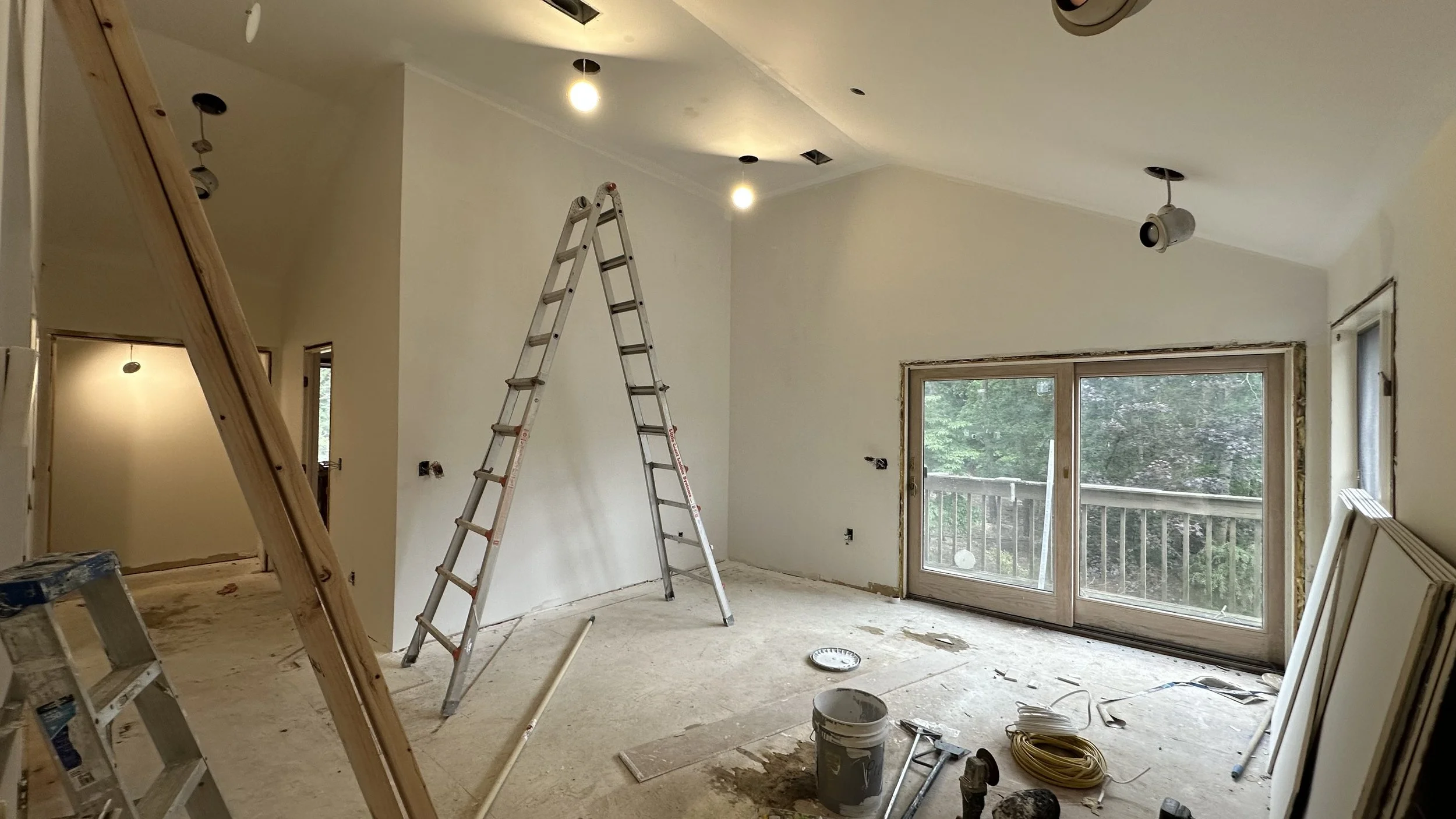 Interior of a room under renovation with construction tools, ladders, a sliding glass door leading to a balcony, and unfinished walls.