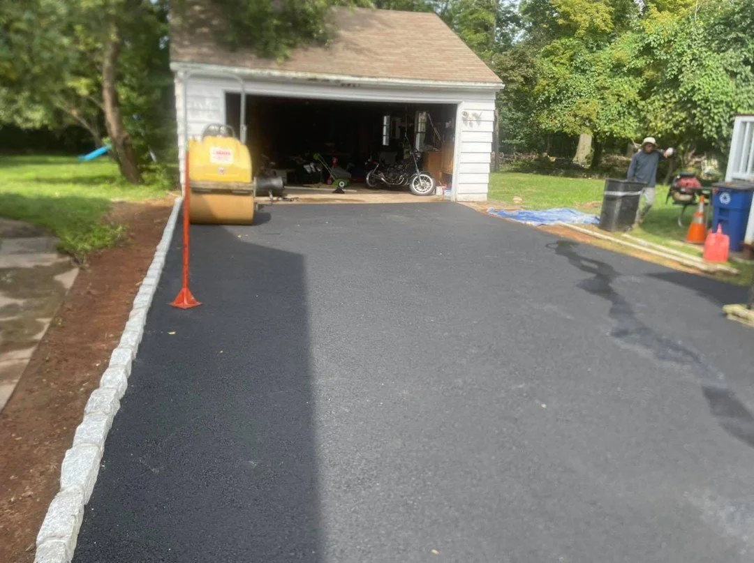 Recently paved driveway in front of a garage, with construction cone and equipment nearby, and a person standing in the yard.