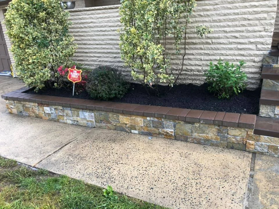 A small landscaped garden bed with three bushes planted in black soil, bordered by a stone wall with various shades of beige, gray, and brown stones, and a row of brown bricks on top. There is a sidewalk in front and steps on the right side of the ga