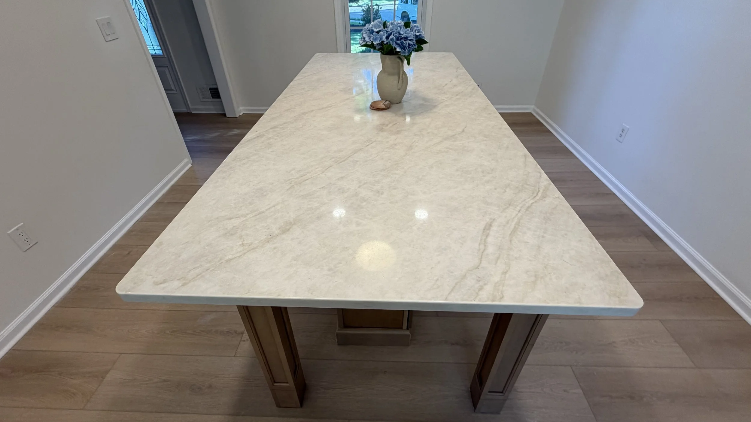 A large rectangular kitchen island with a light-colored marble countertop and wooden support legs in a bright room with hardwood floors and a window behind it, with a vase of blue hydrangea flowers on top.