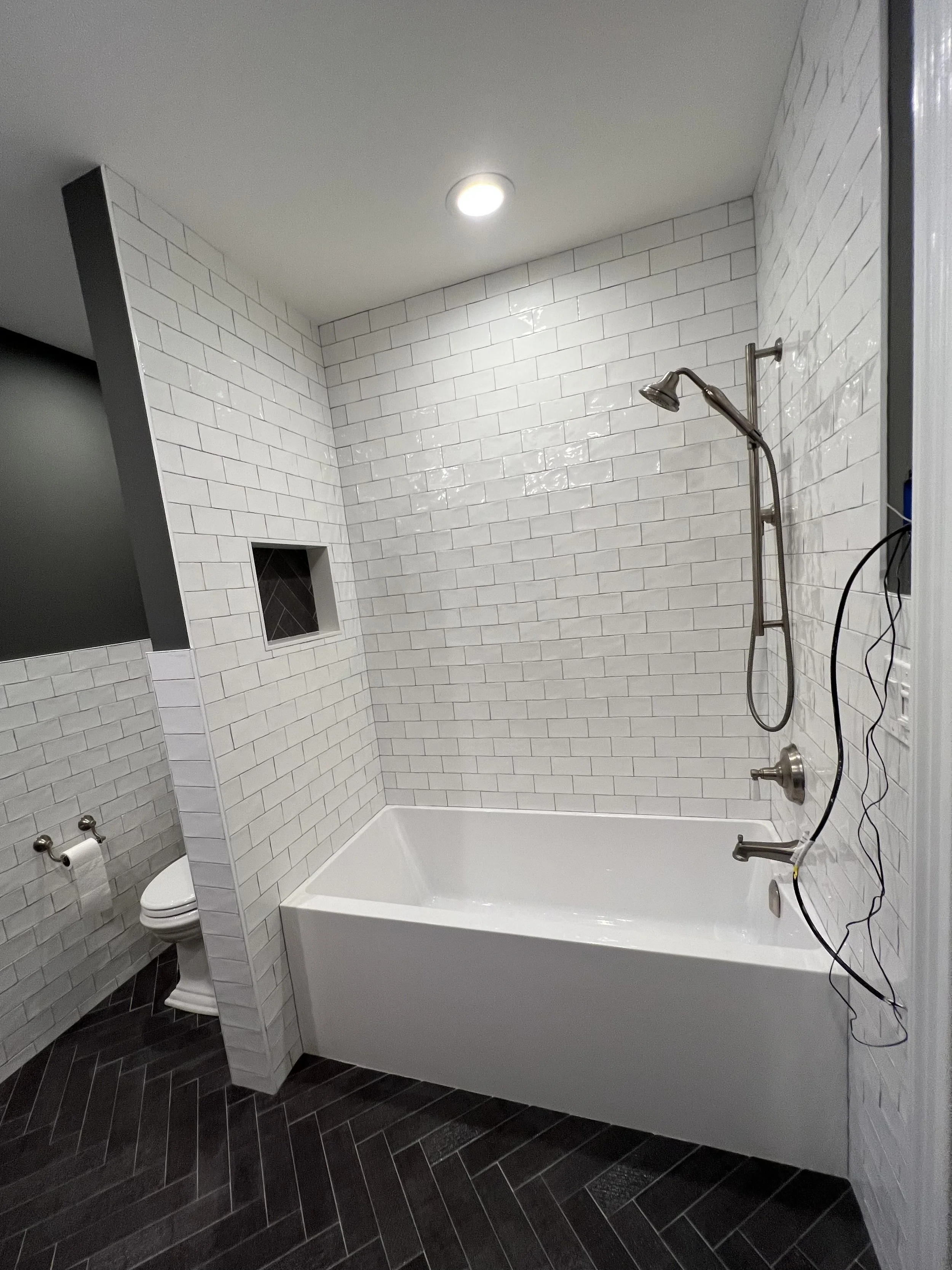 A bathroom with white subway tile walls, a white bathtub, a showerhead, and a black herringbone tile floor. There is a partial view of a toilet and two rolls of toilet paper on a holder. Electrical wires hang from the shower fixtures.
