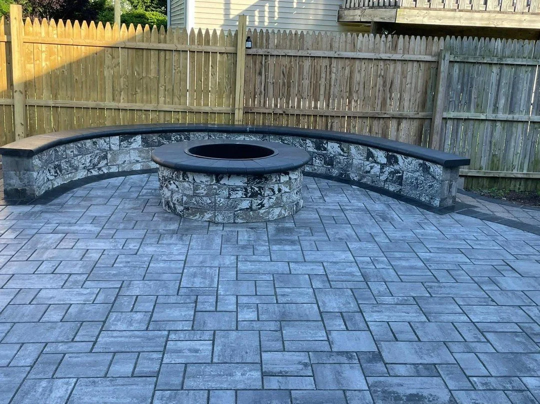 A backyard patio with a round stone fire pit at the center, surrounded by a curved stone bench with a black top. The patio is paved with rectangular concrete pavers and enclosed by a wooden fence, with a house and tree partially visible in the backgr