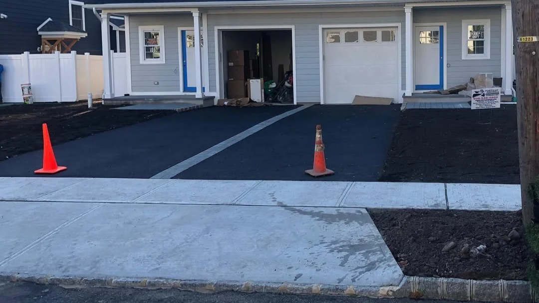 Recently paved driveway leading to a new garage under construction, with orange traffic cones on the sidewalk in front.
