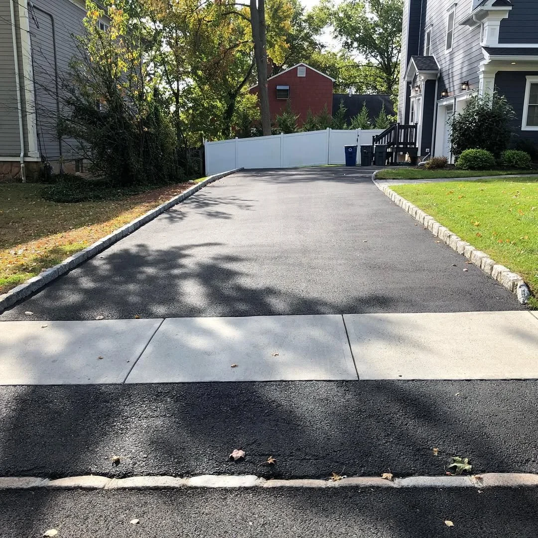 Freshly paved black asphalt driveway leading up to a white fence gate, with a sidewalk and curb in the foreground; residential houses and trees are visible in the background.