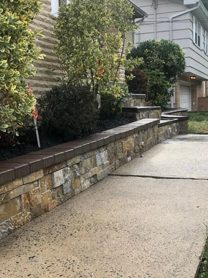 Concrete sidewalk in front of a stone retaining wall with shrubs and plants, house with a garage in the background.