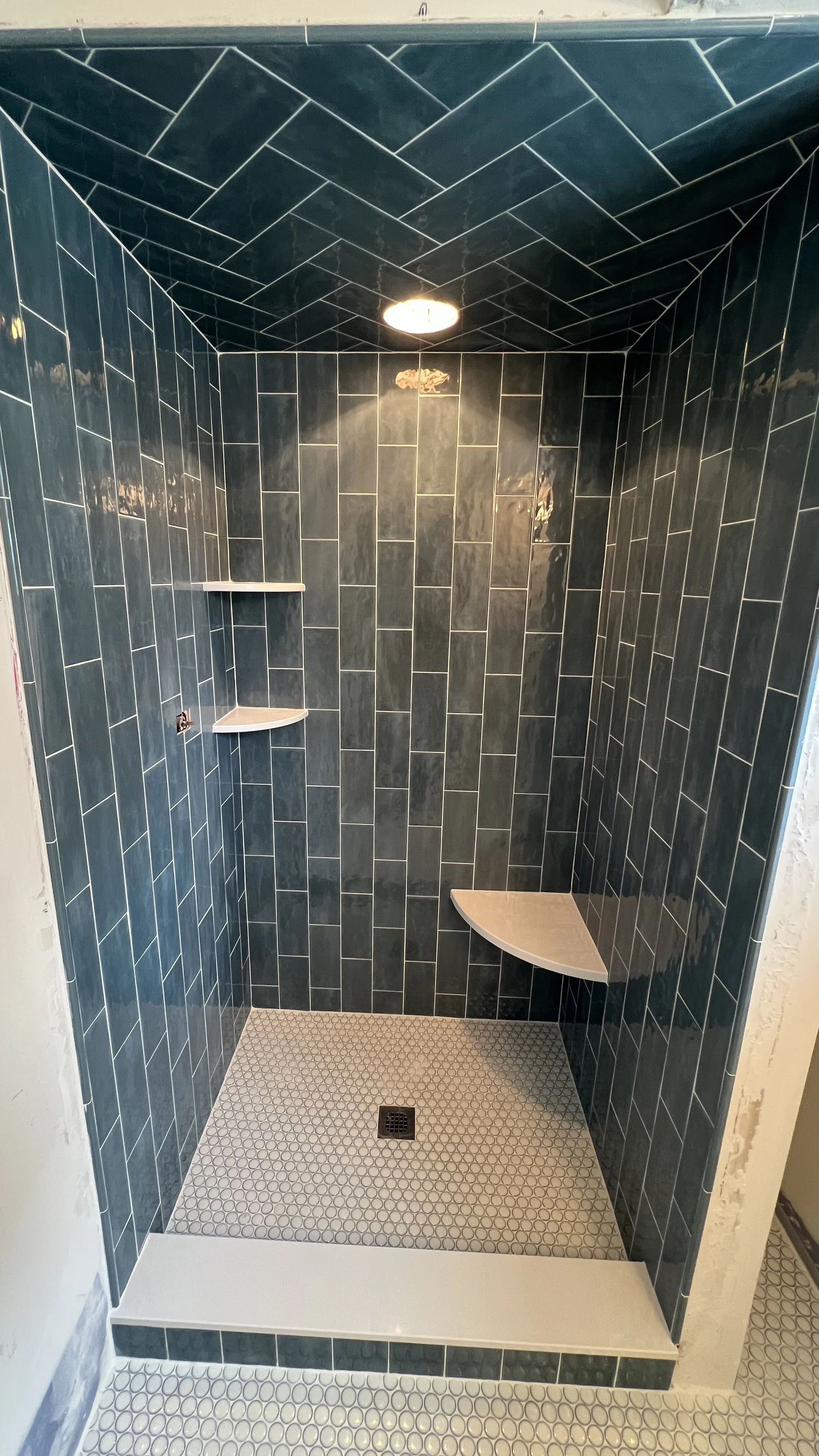 A walk-in shower with dark green tiles on the walls and ceiling, two built-in corner shelves, beige bench seat, small square beige tiles on the floor, and a central drain.