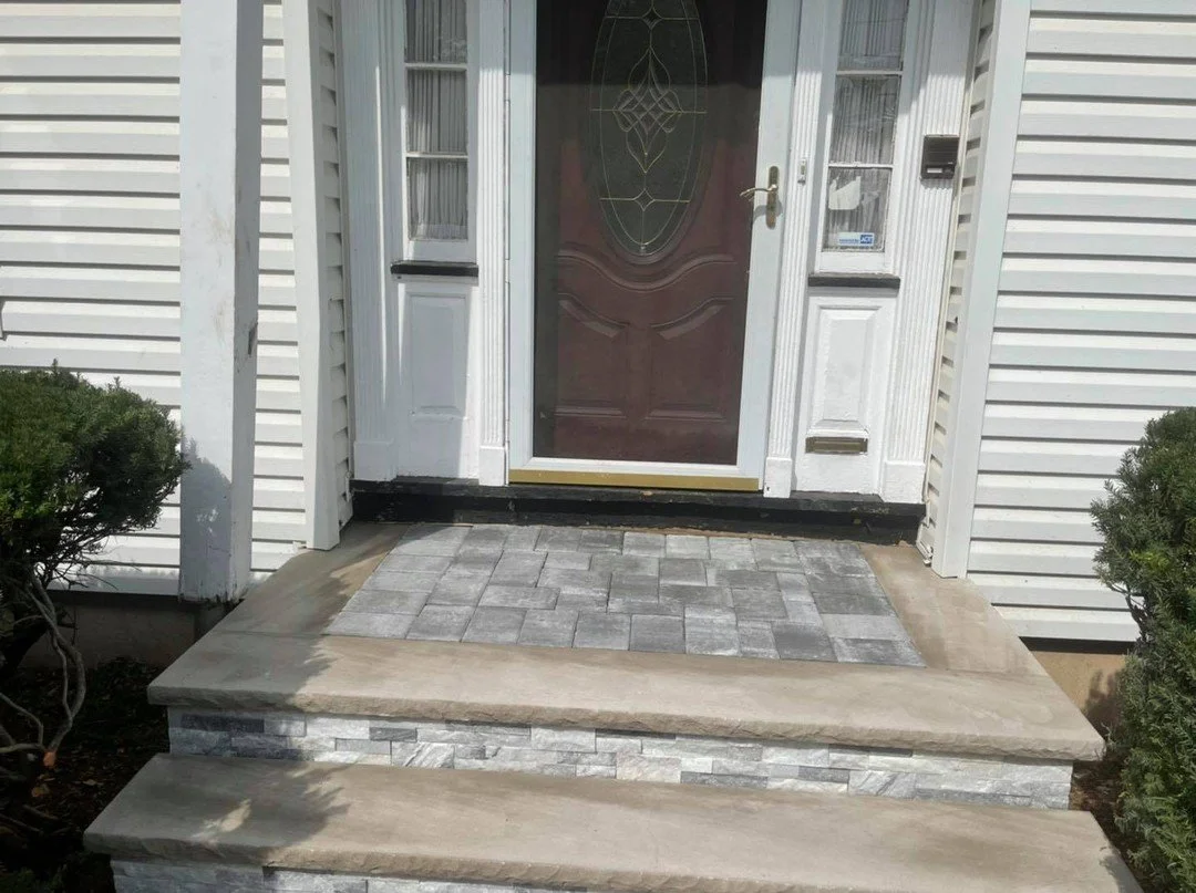 Front porch with gray brick steps, a brown door with decorative glass, white trim, and two small bushes on each side of the stairs.