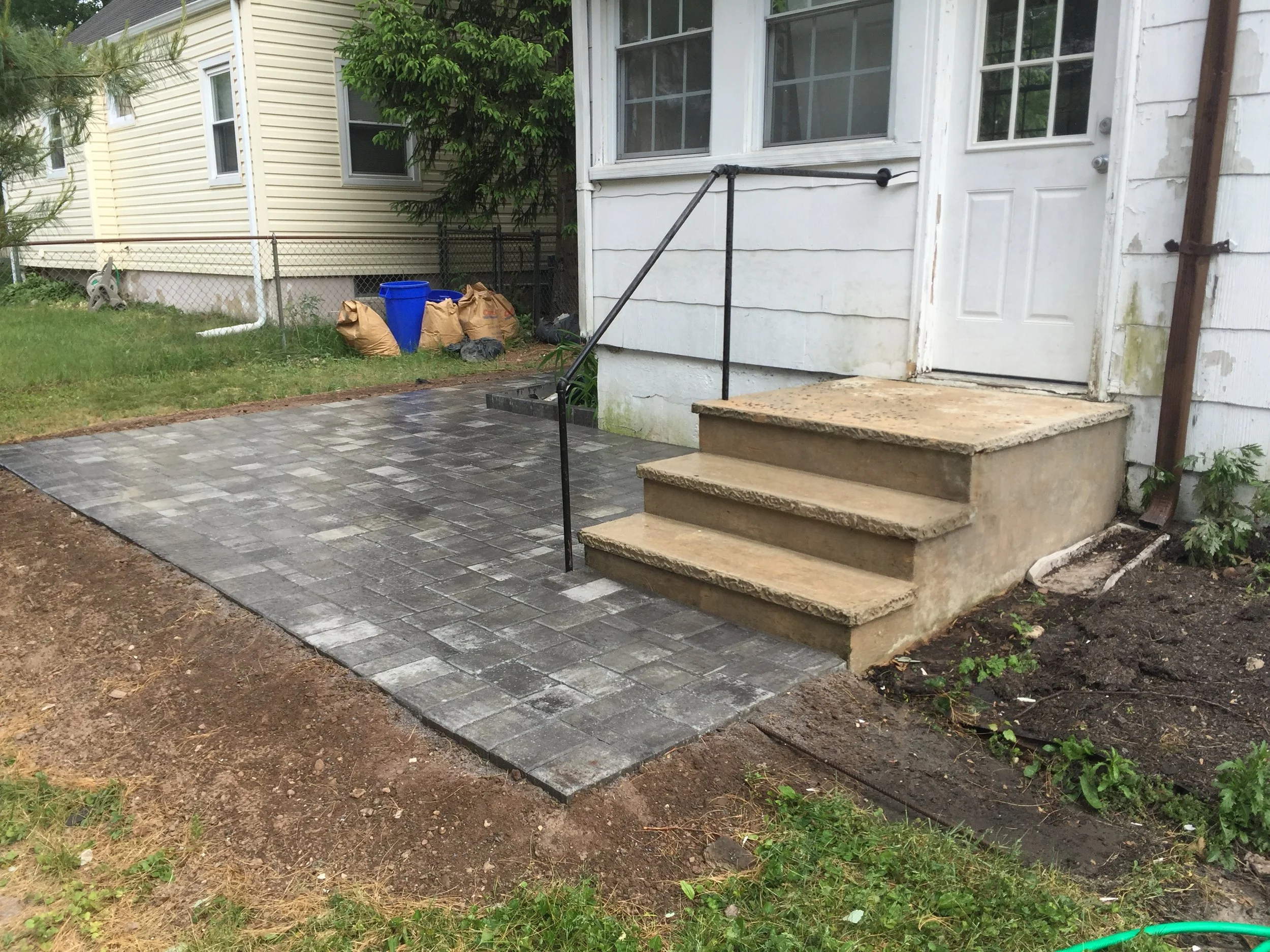 Newly finished brick patio with stairs and black handrail leading to the white door of a house. Patches of dirt and grass around the patio, with construction supplies in the background.