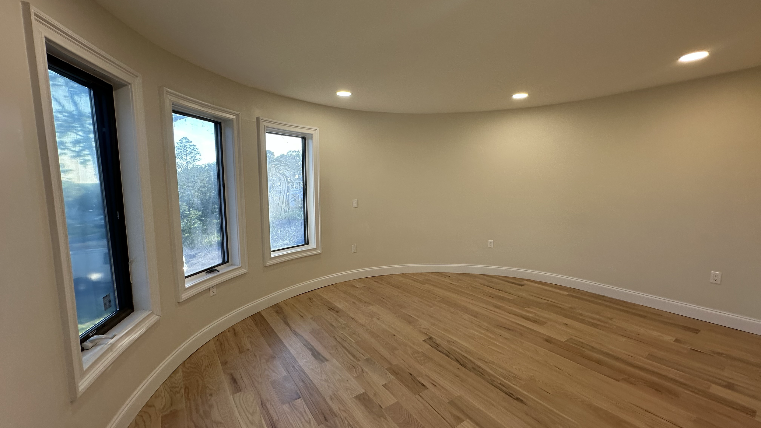 Empty room with hardwood floors, beige walls, and three large windows with a view of trees outside. Recessed ceiling lights are installed.