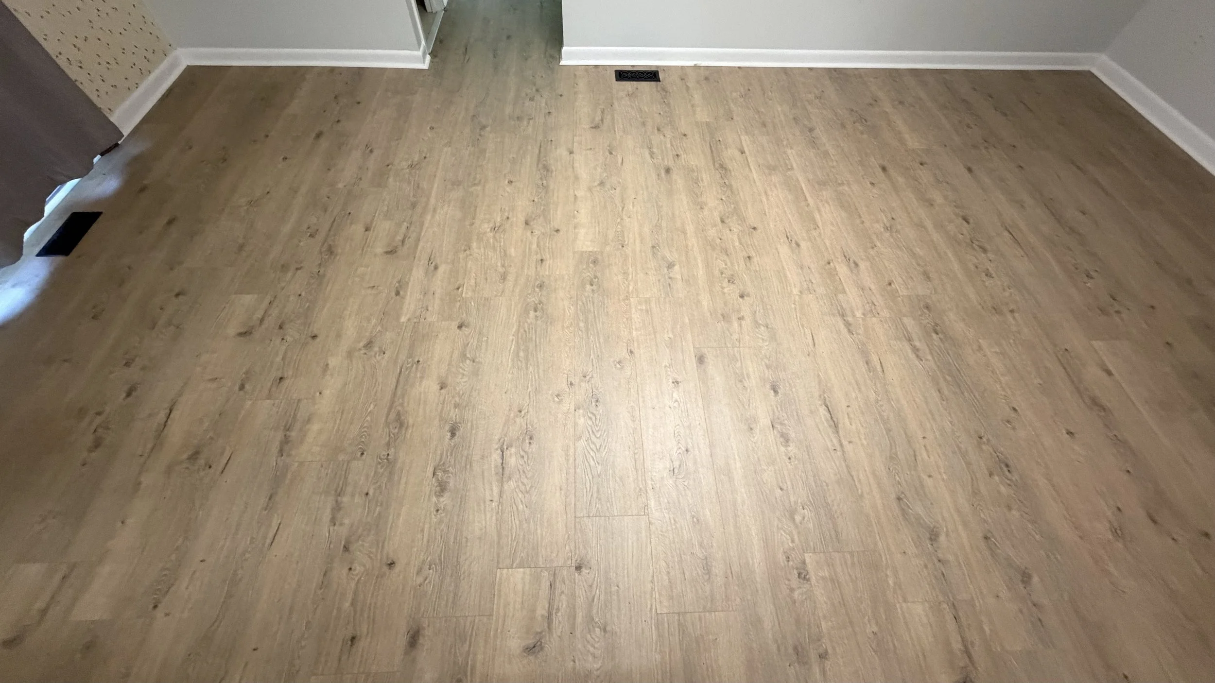 A room with light brown wood flooring, baseboards, and white walls. There are vents on the floor and a partial view of a doorway and wall with different textures.