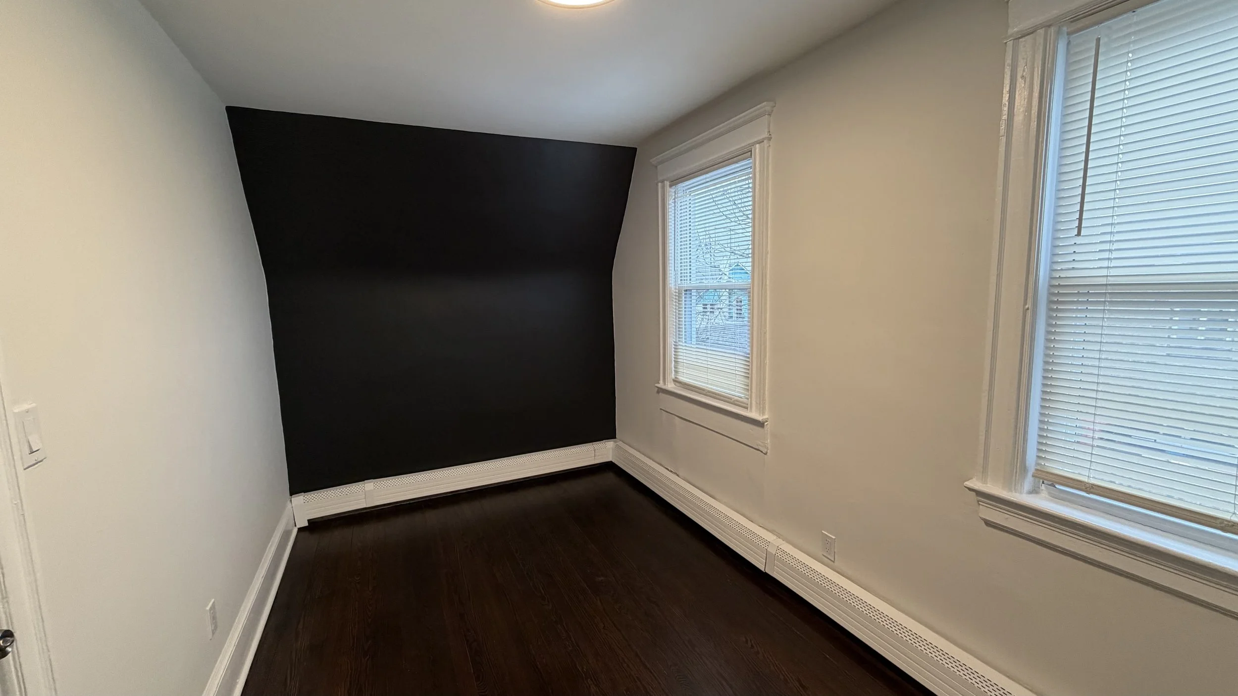 Empty room with two windows with blinds and a black accent wall on the left side. White walls and trim, dark wood floor, baseboard heating vent along the bottom of the walls.