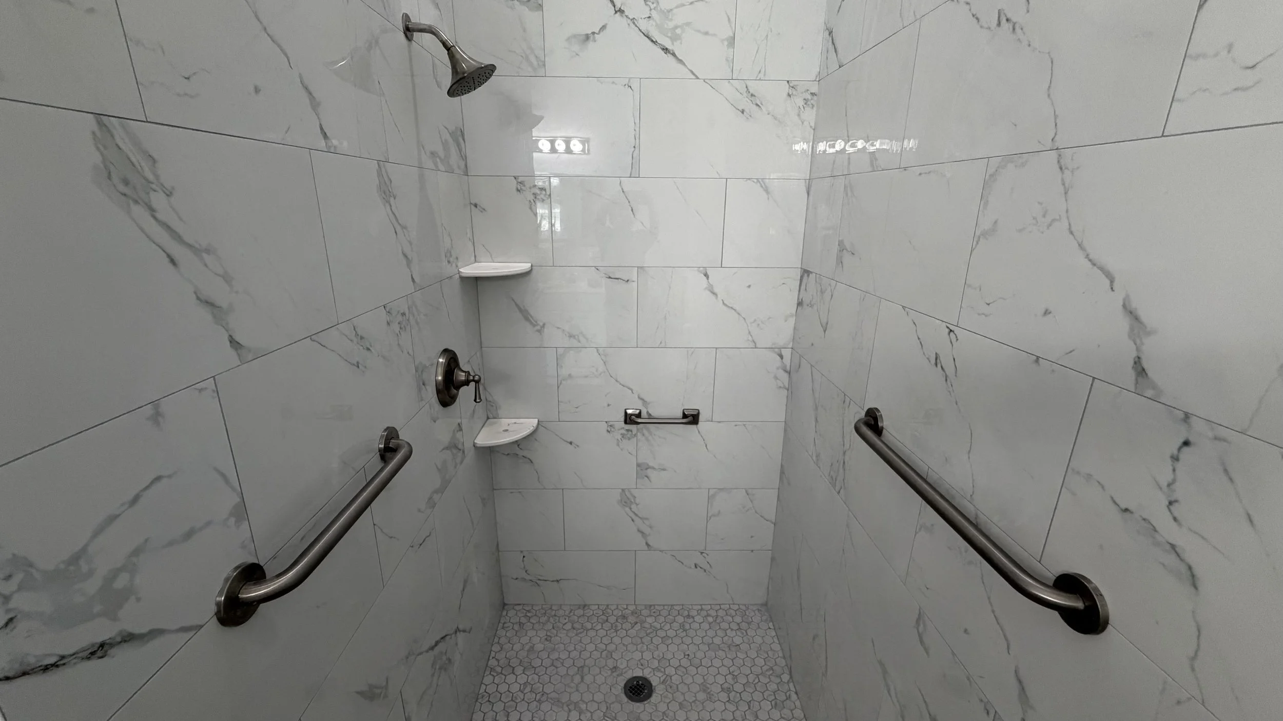 A walk-in shower with marble walls, a handheld showerhead, a shelf, grab bars, and small hexagonal tiles on the floor.