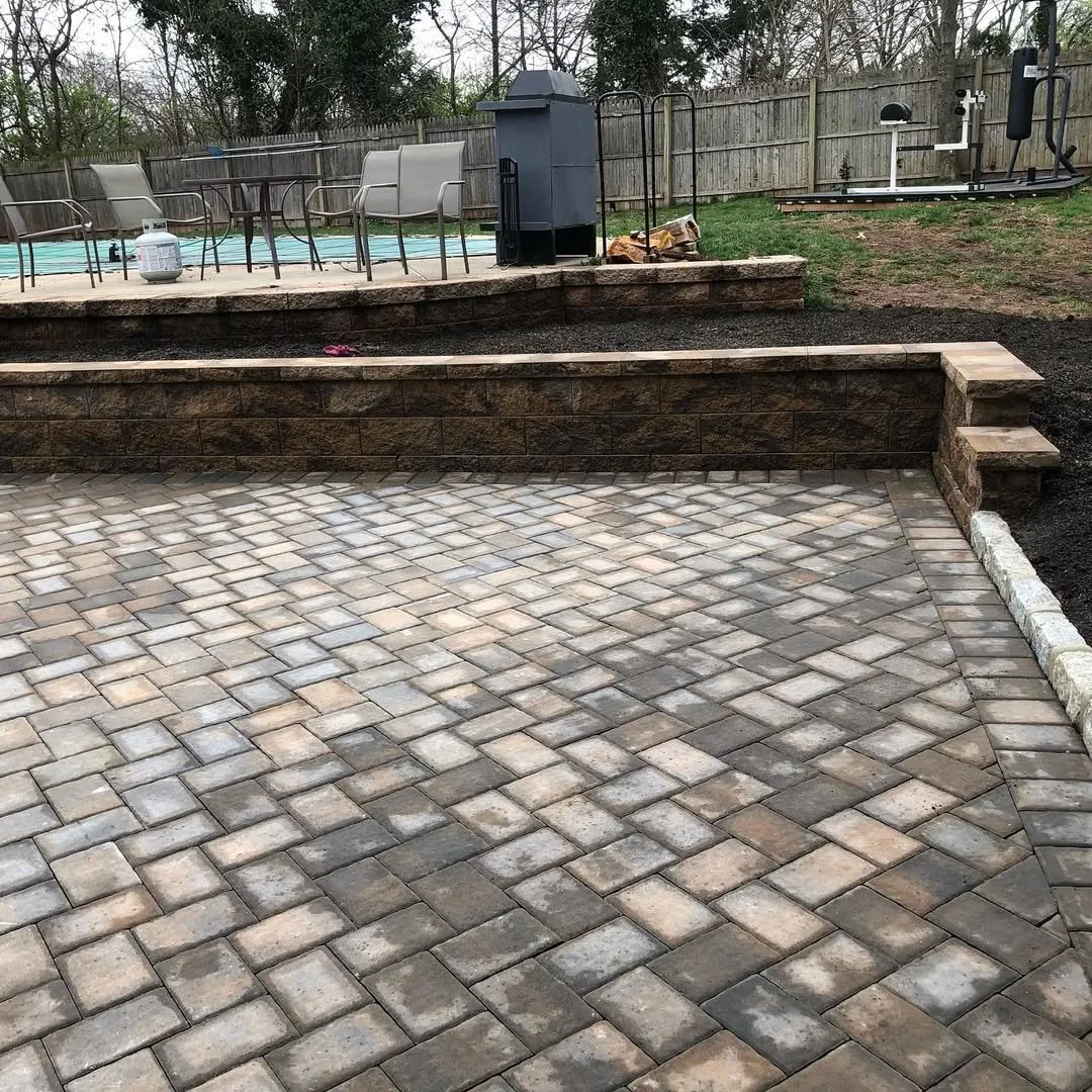 Backyard patio with brick pavers, low retaining wall, outdoor furniture, grill, and a fenced yard with some trees.