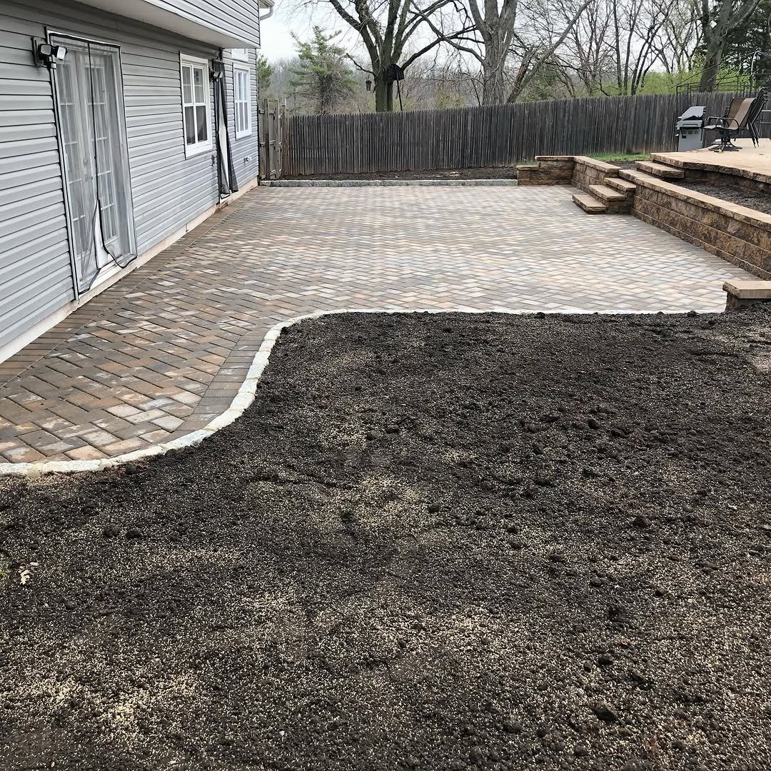 A backyard with a partially completed paved patio and stairs, with a section of dark dirt or gravel in the foreground, and a house with siding and sliding glass doors on the left side. There is a wooden fence and some outdoor chairs with a grill in t