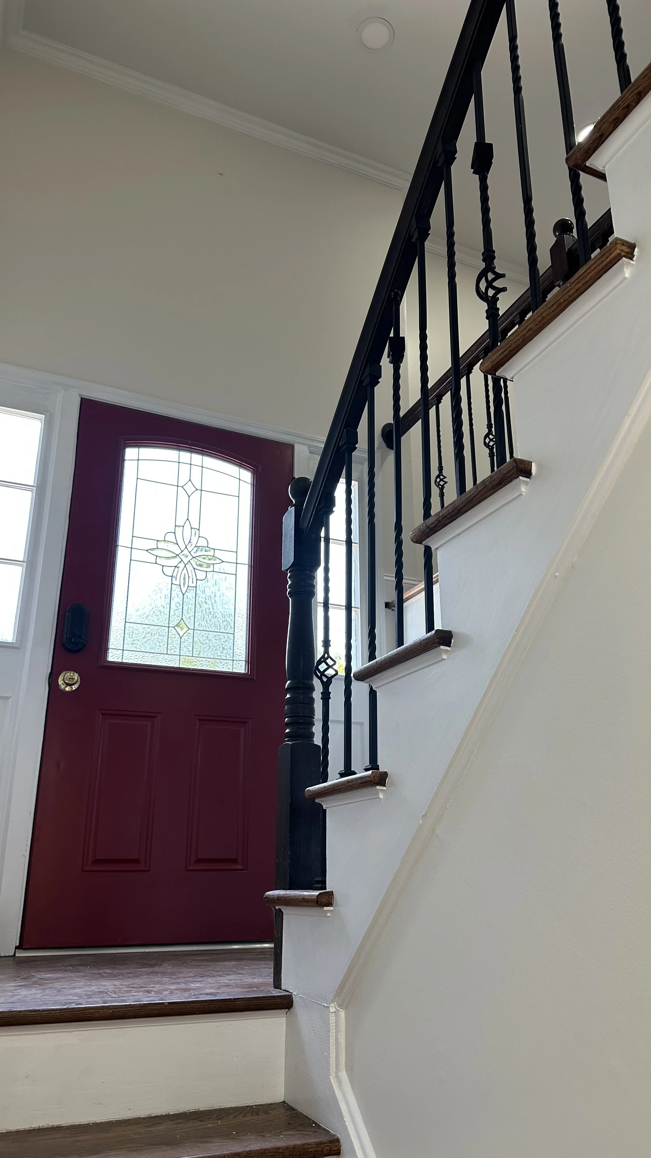 Interior view of a home's front entryway with a red front door featuring decorative glass, a black railing staircase, and a wooden staircase.