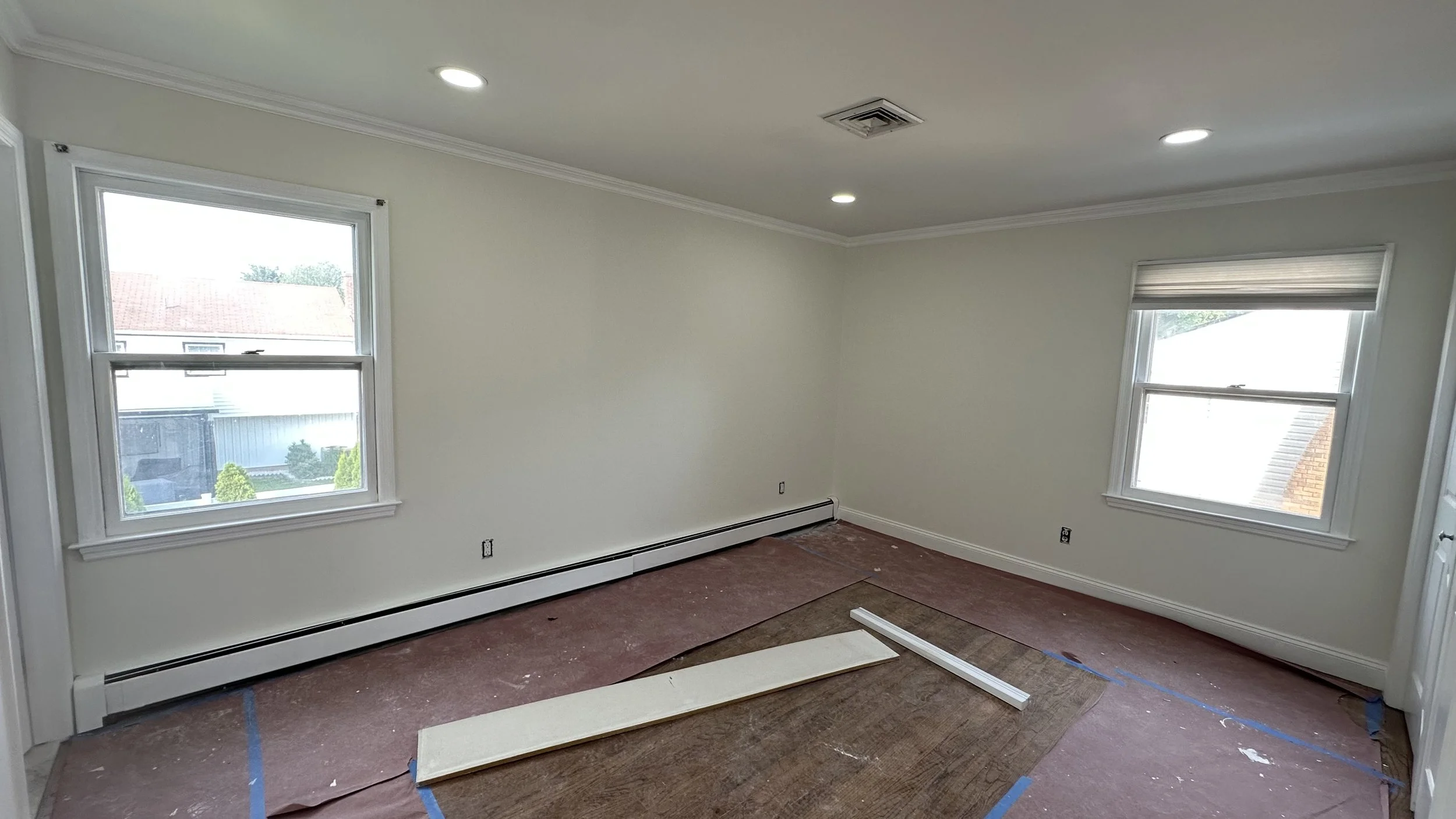 Empty room with two windows, white walls, crown molding, ceiling lights, and a cap heater along the baseboard, with flooring under renovation and protective covering on the floor.