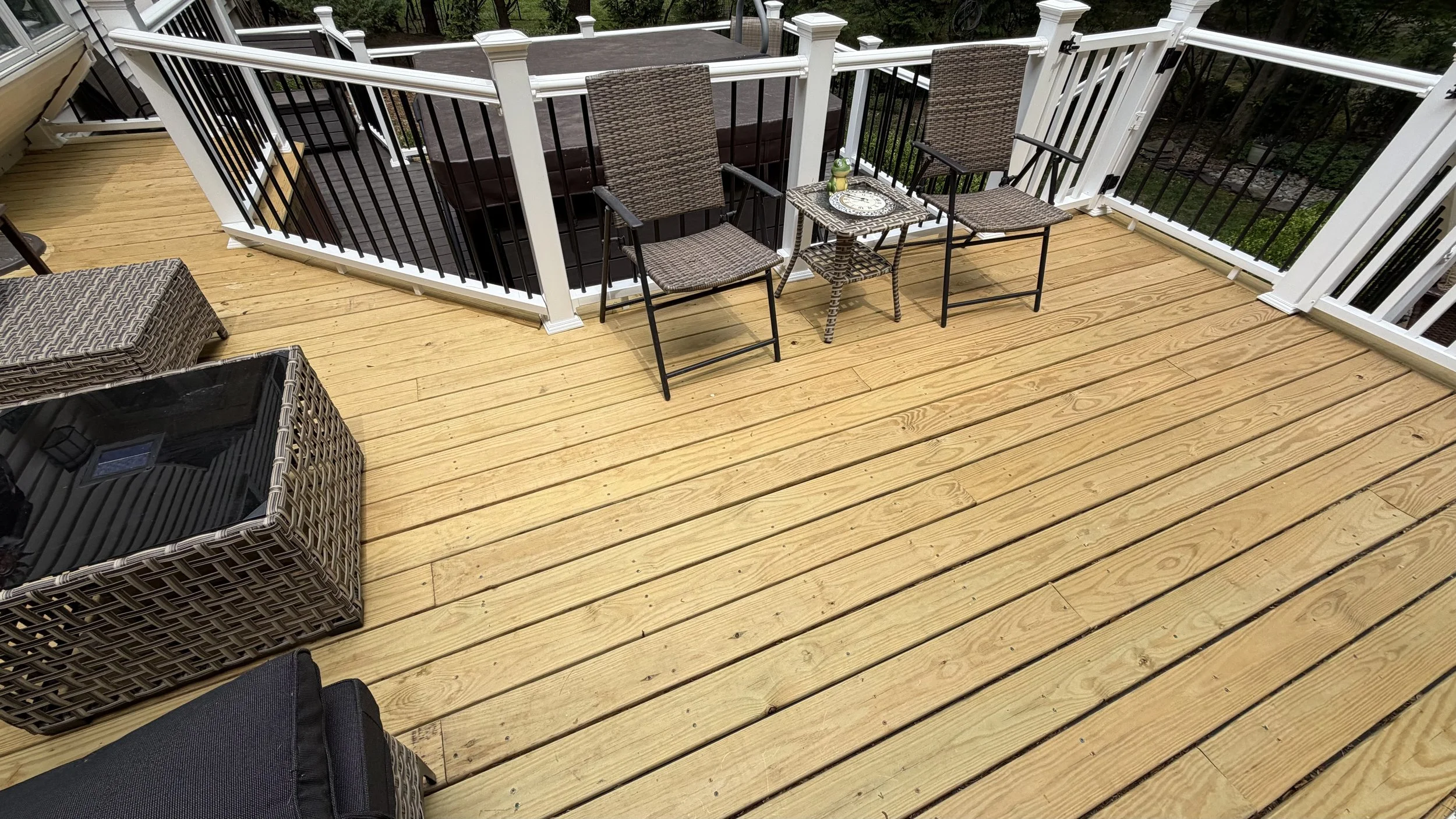 View of a wooden deck with black, brown, and beige outdoor furniture, including chairs and tables, with railing and stairs leading down to a green yard.
