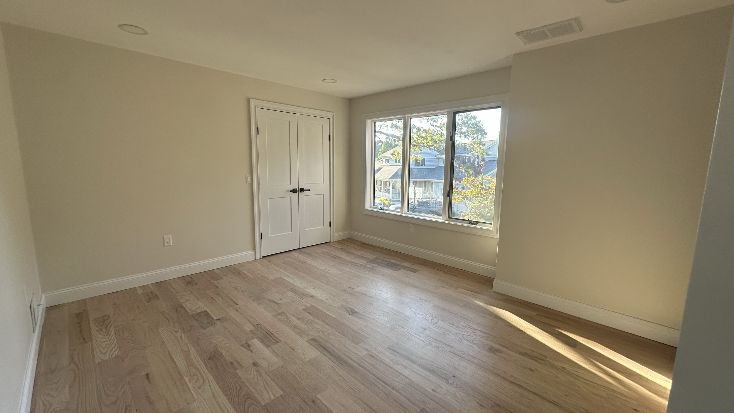 Empty room with light wood floors, white walls, a window showing a neighborhood view, and a closed double door